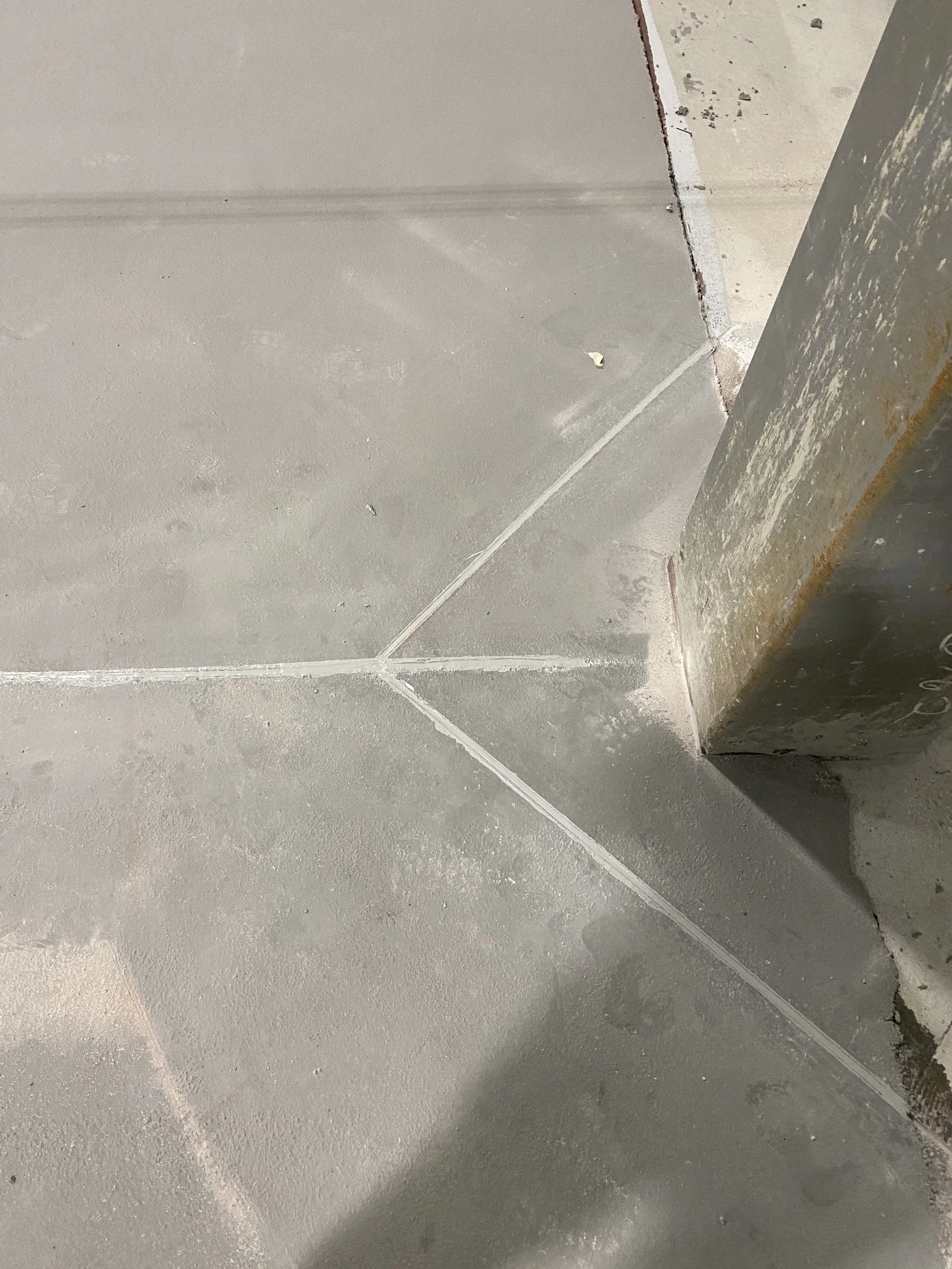 Close-up of concrete floor with grout lines at the corner of a room, showing part of a weathered wall or column.