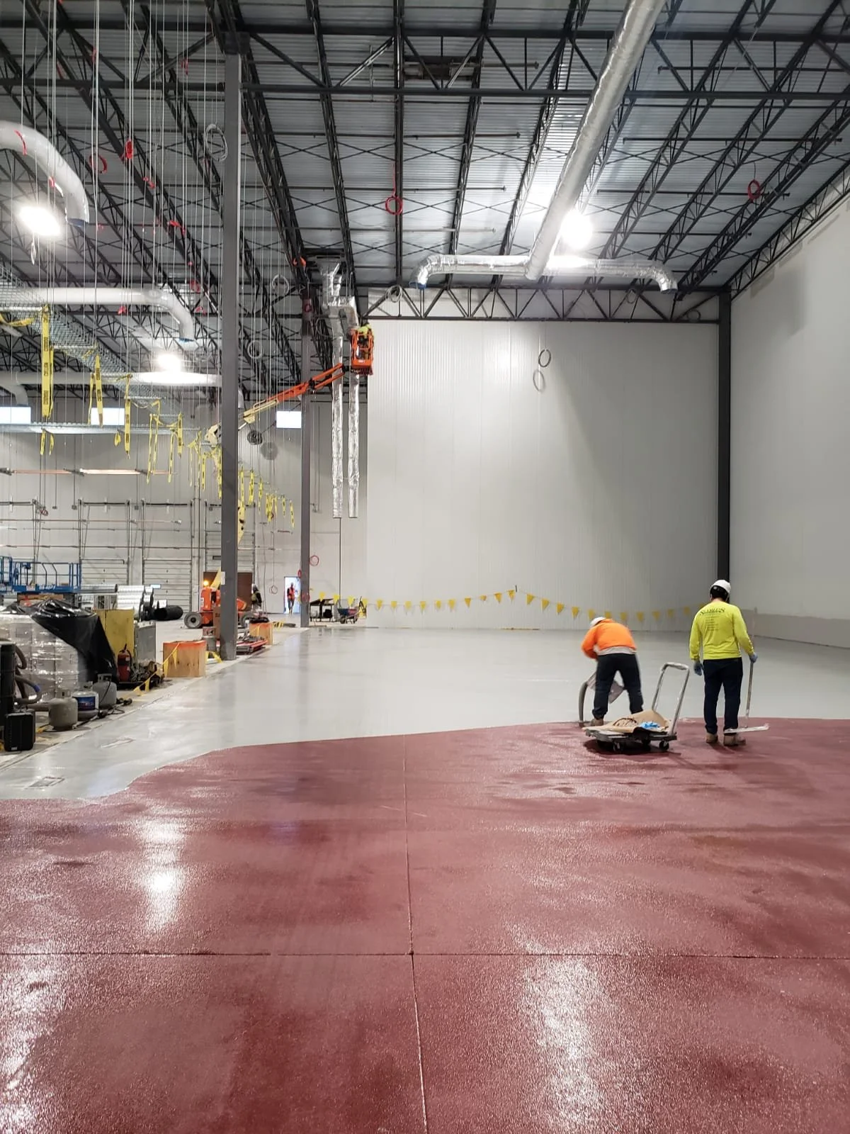 Construction workers installing flooring in a large industrial warehouse with metal beams and ductwork ceiling.