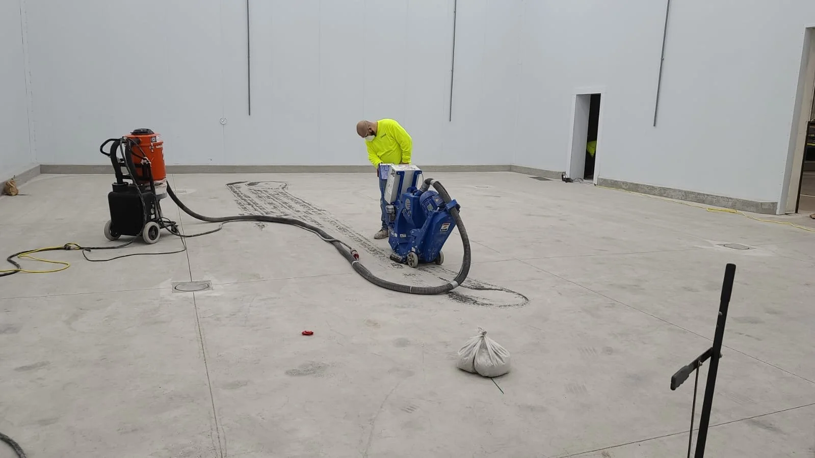 Construction worker in a yellow shirt operating concrete floor grinding machines in an unfinished indoor space.