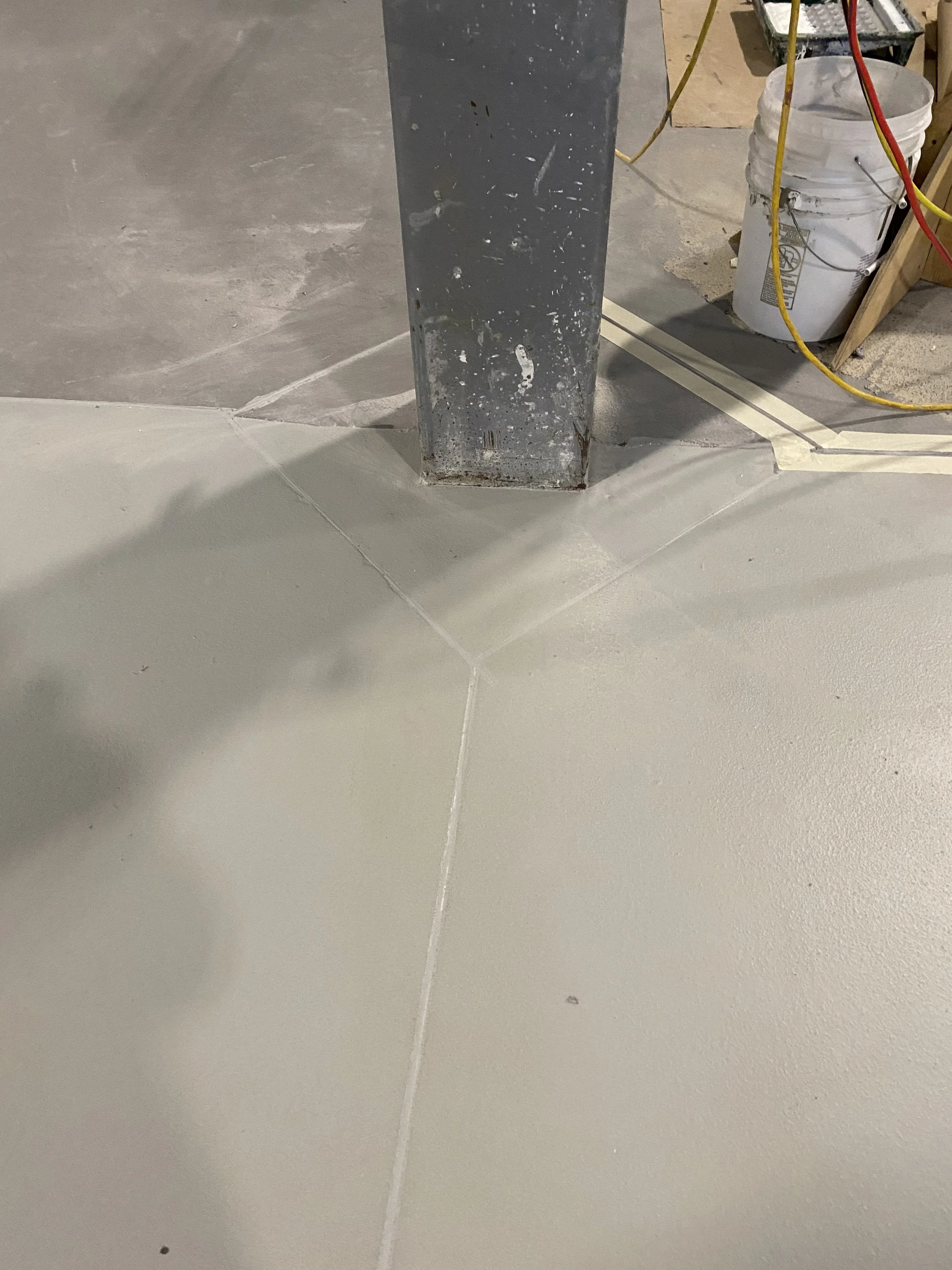 Close-up of a metal support beam at the intersection of concrete floor tiles in a construction site. The beam shows signs of rust and paint chipping, with construction materials and equipment in the background.