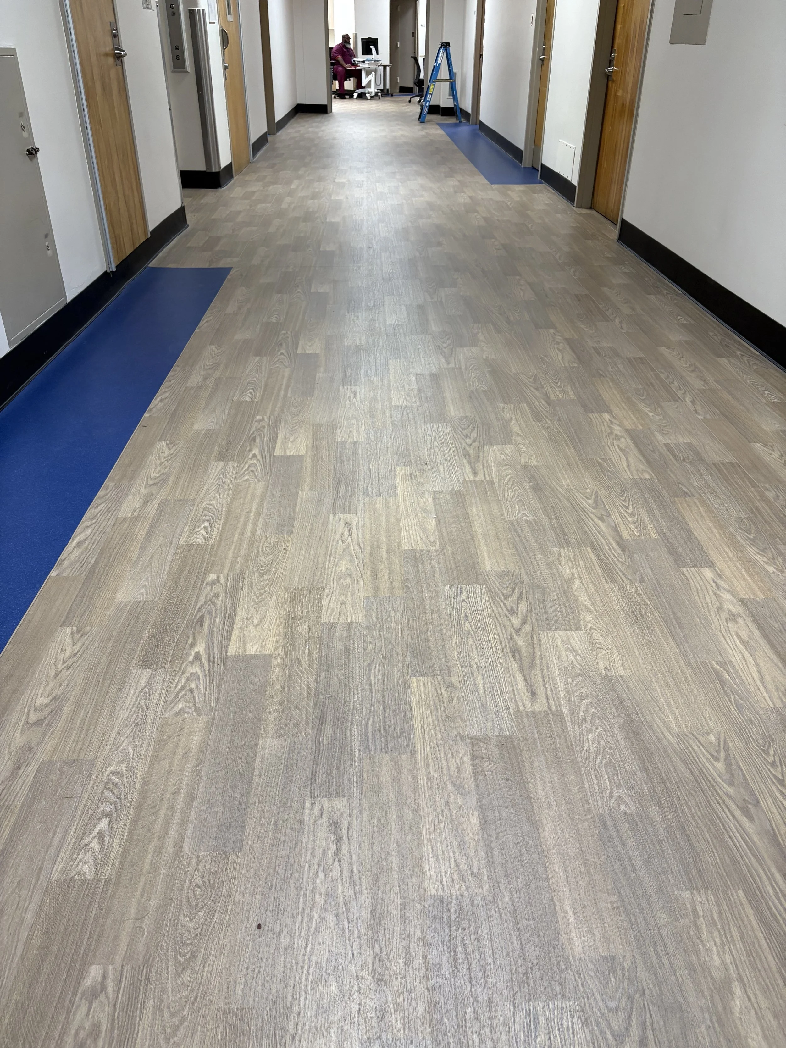 Empty hospital hallway with wood-look flooring, white walls, wooden doors, blue mats, and a person working at a desk in the background.