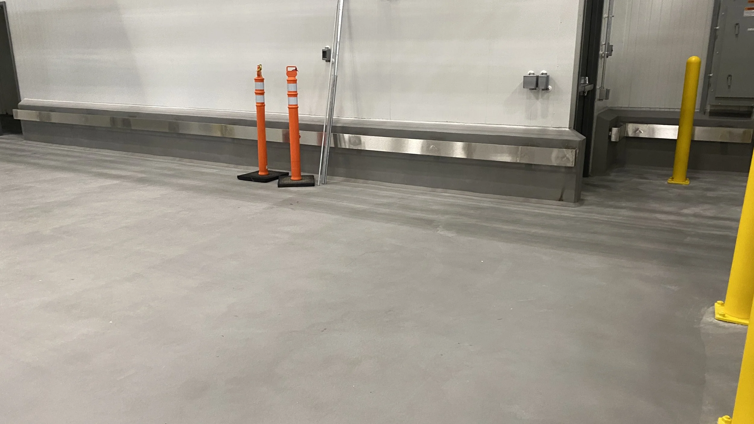 Empty industrial or warehouse floor with a large, white insulated door, yellow and orange safety posts, and a metal barrier.