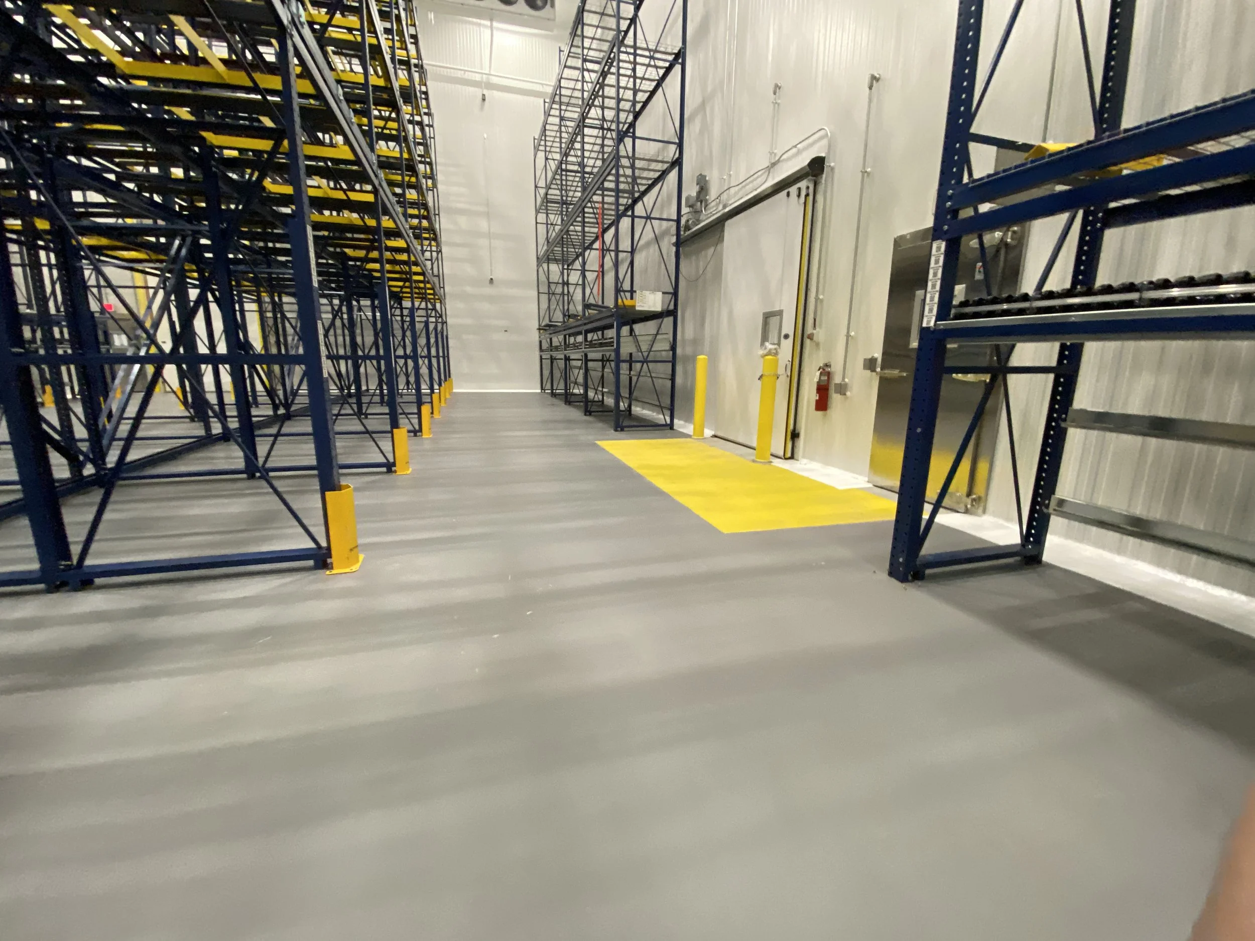 Empty warehouse with metal shelving racks on both sides, yellow caution markings on the floor and safety barriers around electrical panels and fire extinguisher.