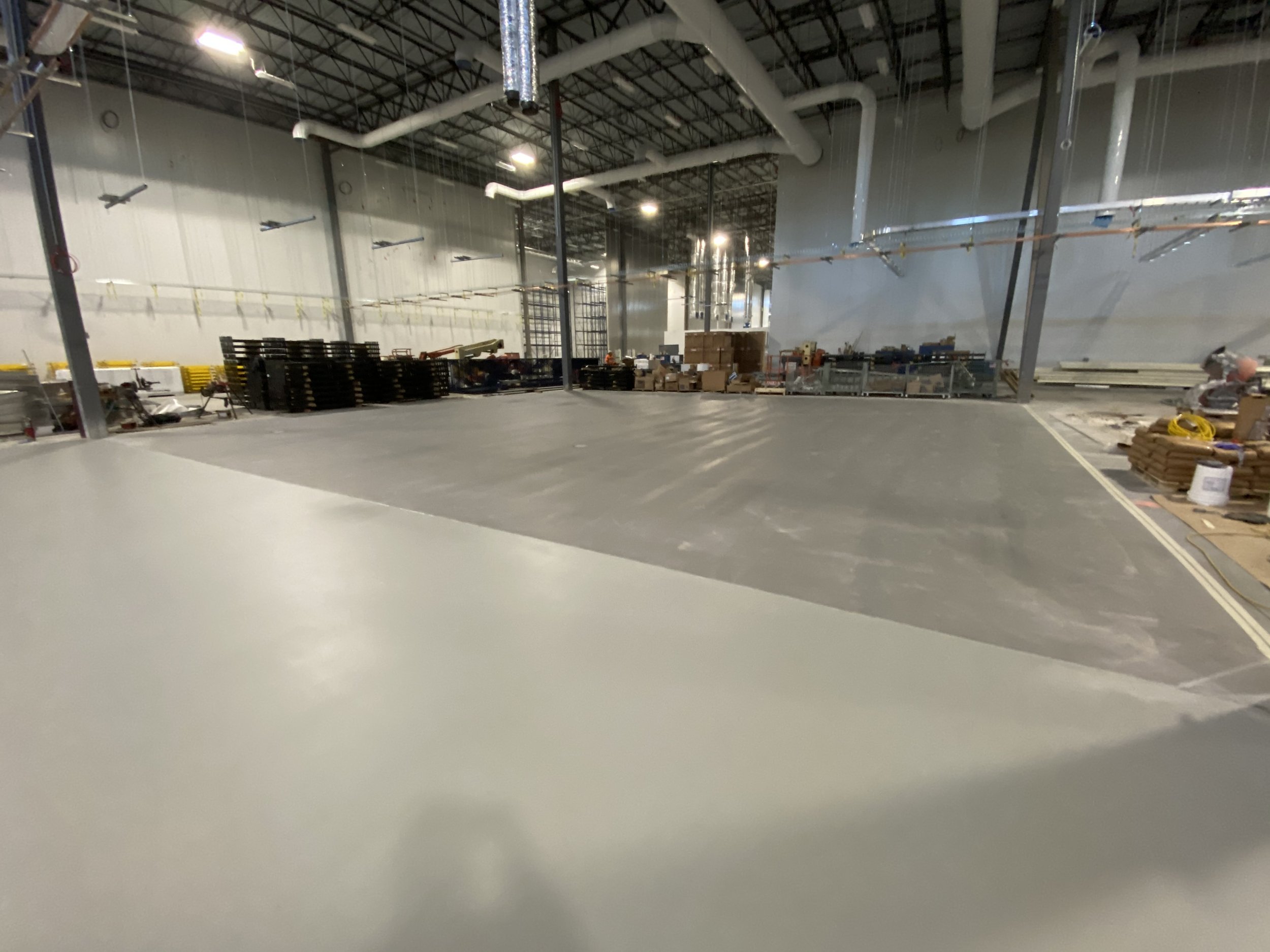 Interior of an unfinished industrial or warehouse space with a concrete floor and exposed ceiling. Construction materials and equipment are stacked along the walls.