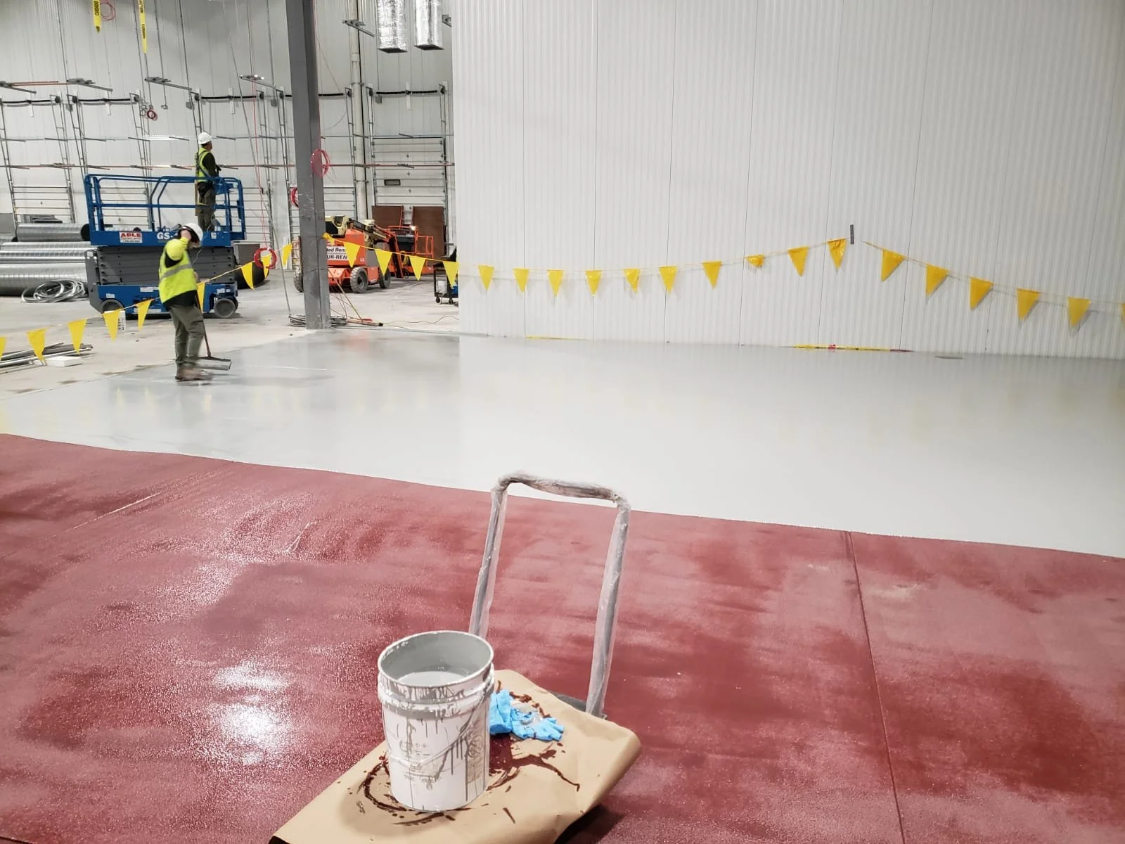 Workers are applying a light gray epoxy coating on the floor of a large industrial space, with one section of the floor already coated in red epoxy. Construction equipment and supplies are visible in the background.