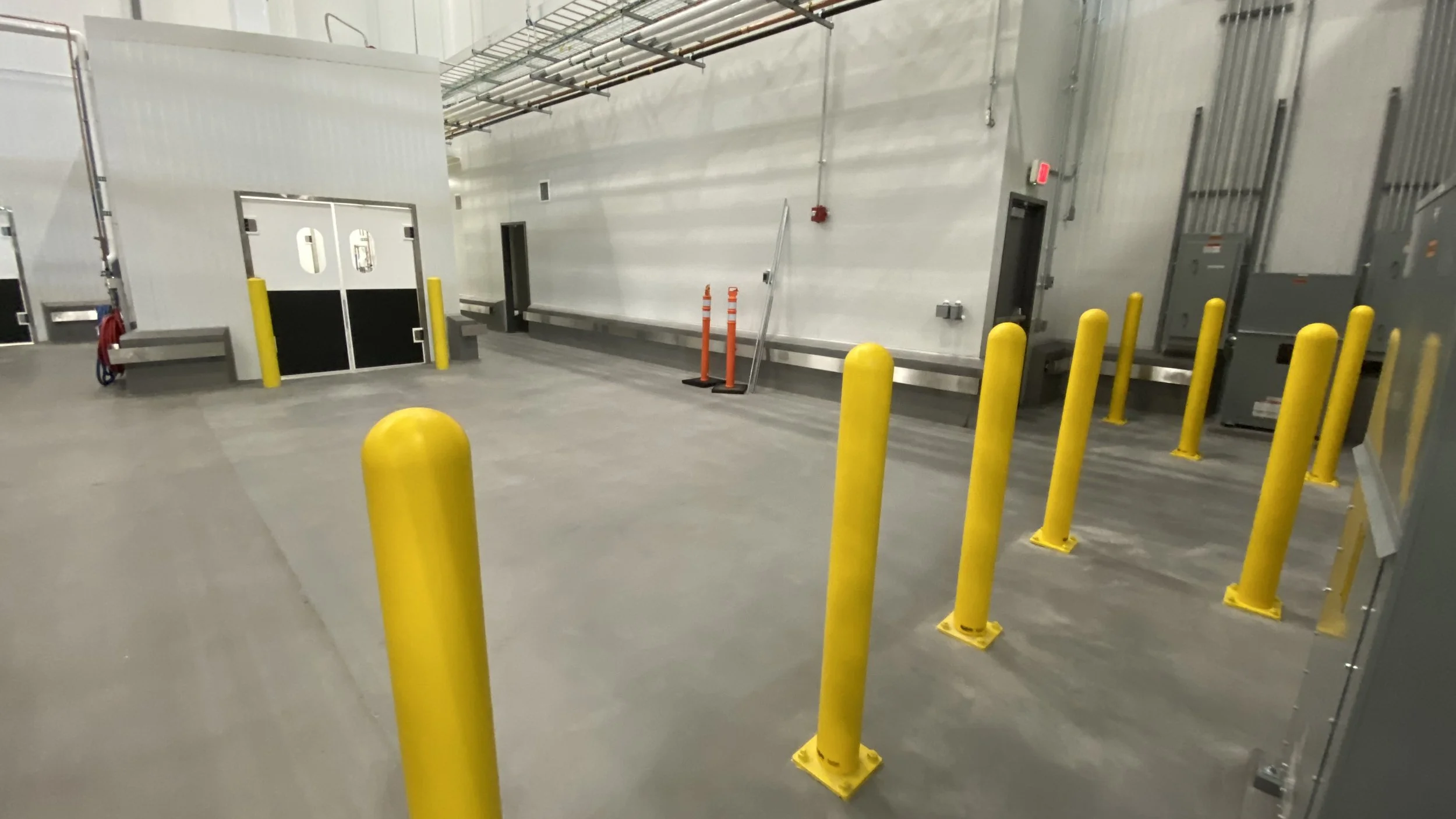 Empty loading dock area with yellow safety bollards, gray concrete floor, and metallic walls with doors and utility panels.