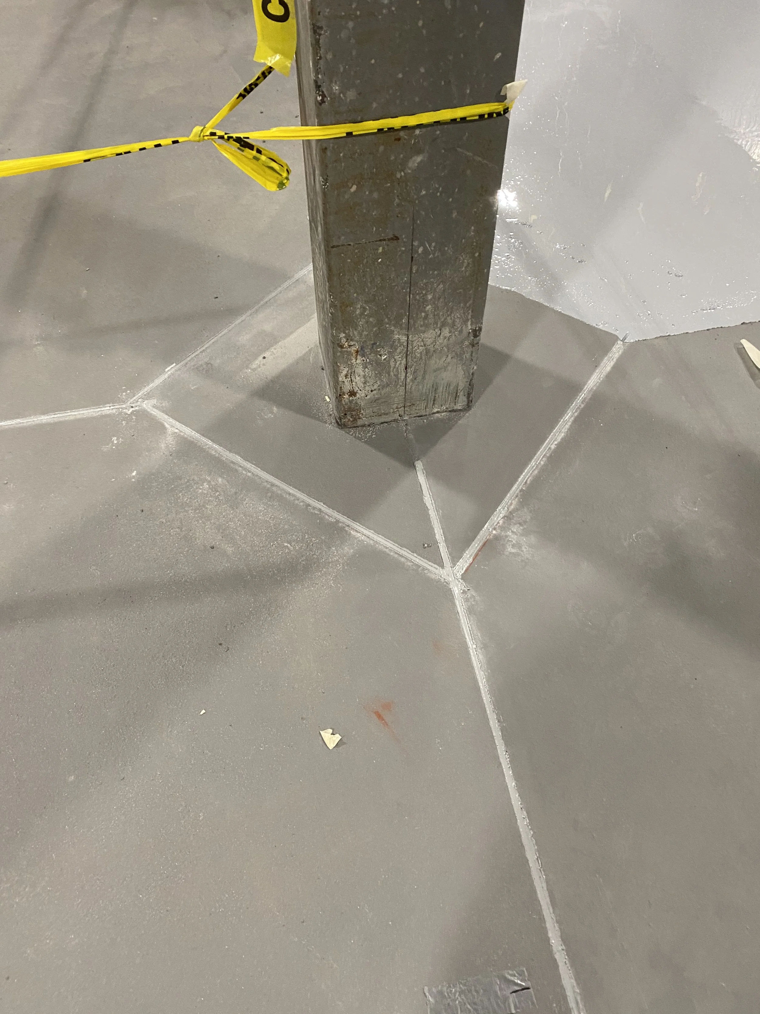 Close-up of a concrete floor with freshly painted or sealed lines in a grid pattern, a metal pole at the corner, and yellow caution tape wrapped around part of the pole.