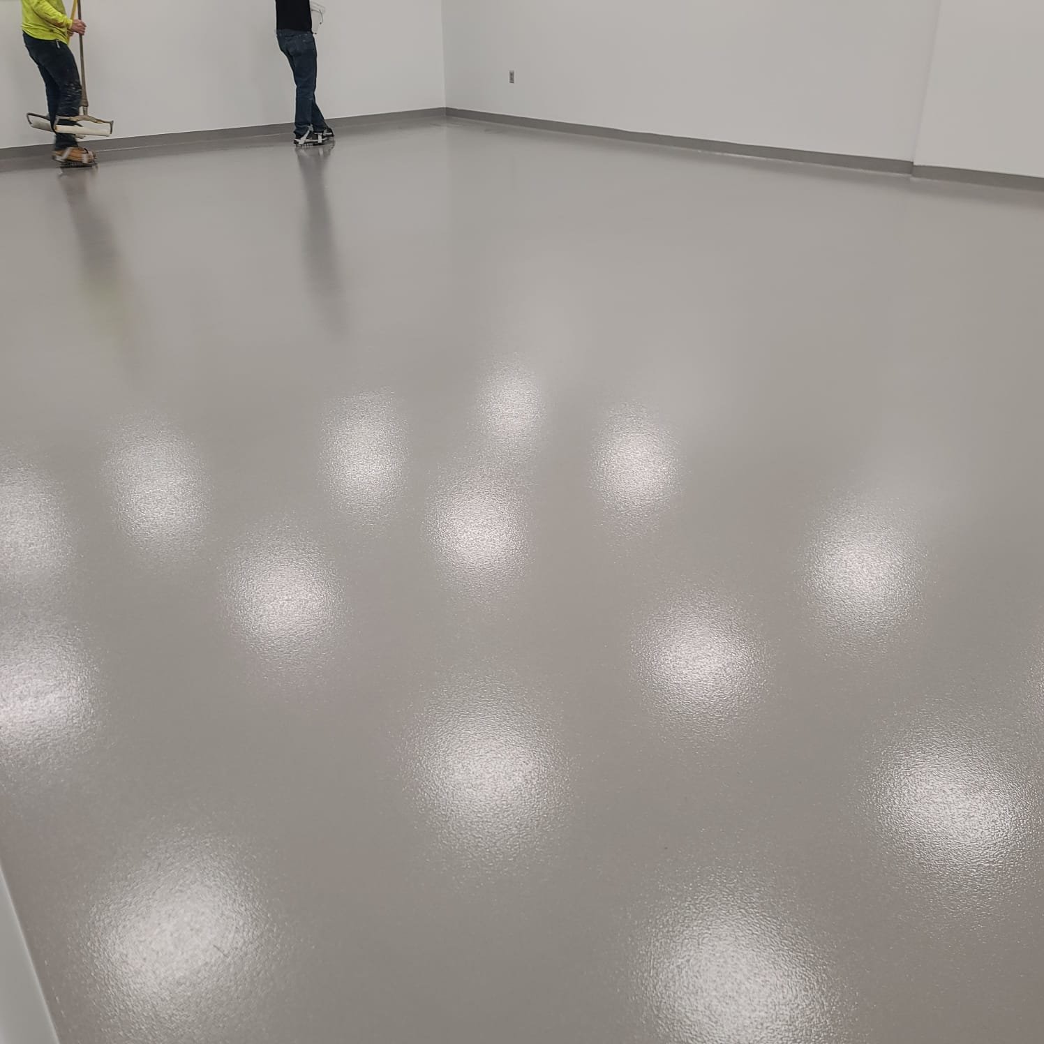 Clean, shiny gray flooring in a large empty room with white walls. Two people are working on the floor, one using a mop and the other on a skateboard.