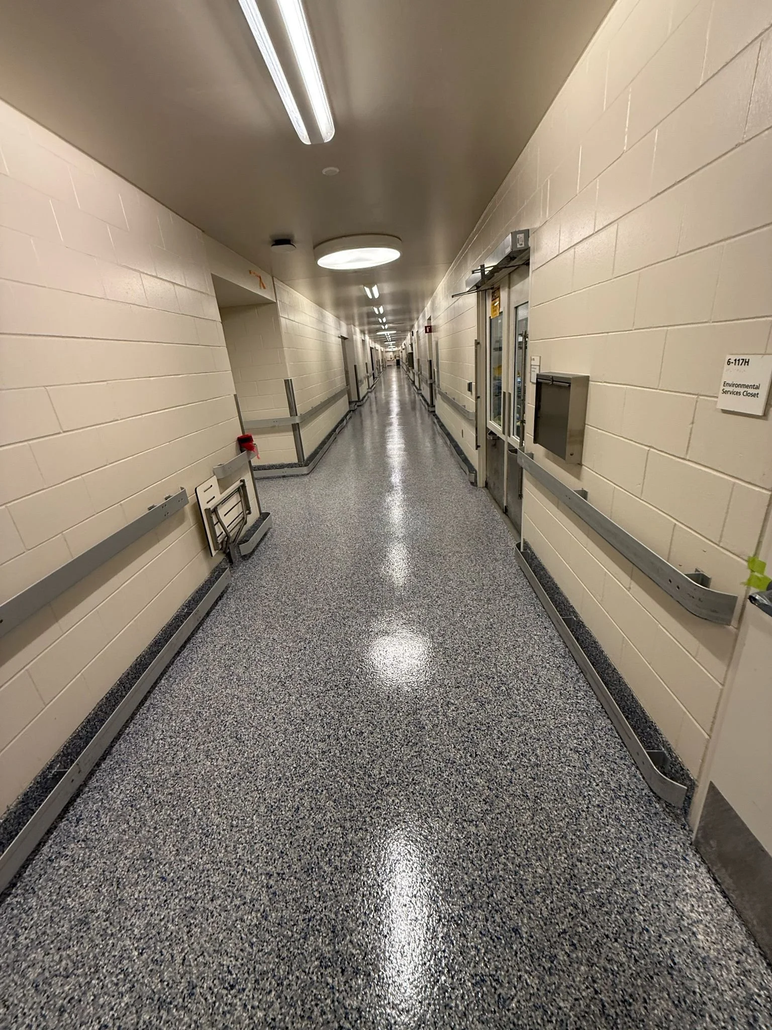 Long hospital corridor with beige brick walls, gray speckled floor, ceiling lights, and a few signs on the wall, including a sign for an environmental services closet.