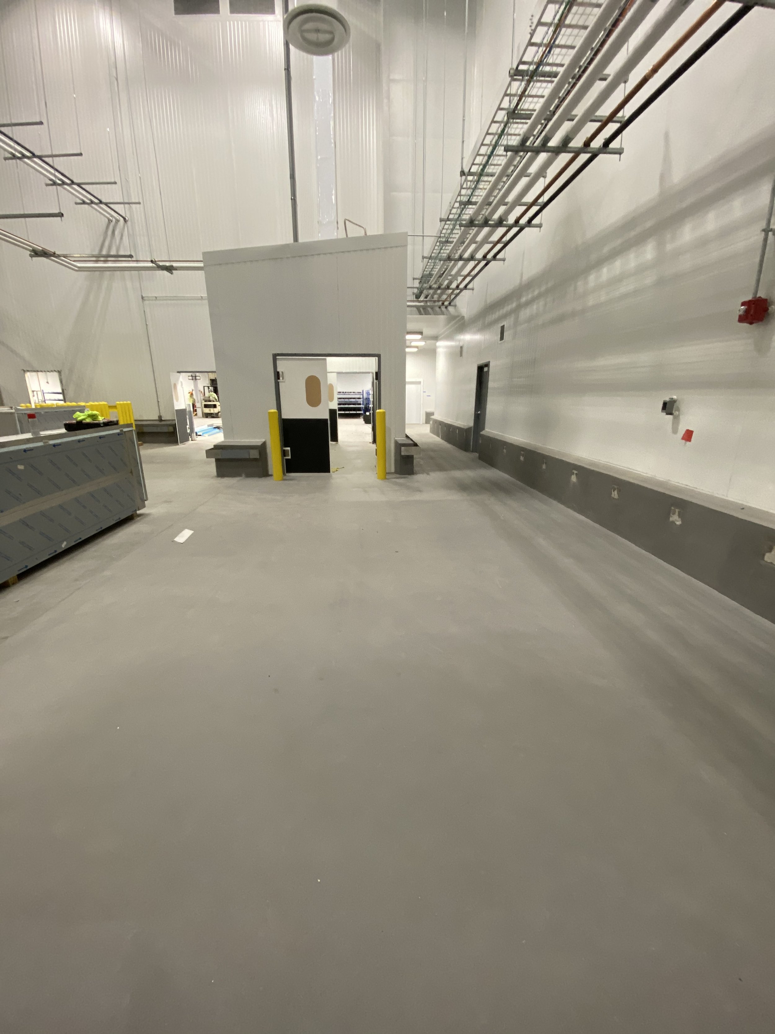 Empty industrial warehouse or factory with high ceiling, white walls, and exposed piping, and an enclosed small room or guard booth in the center.