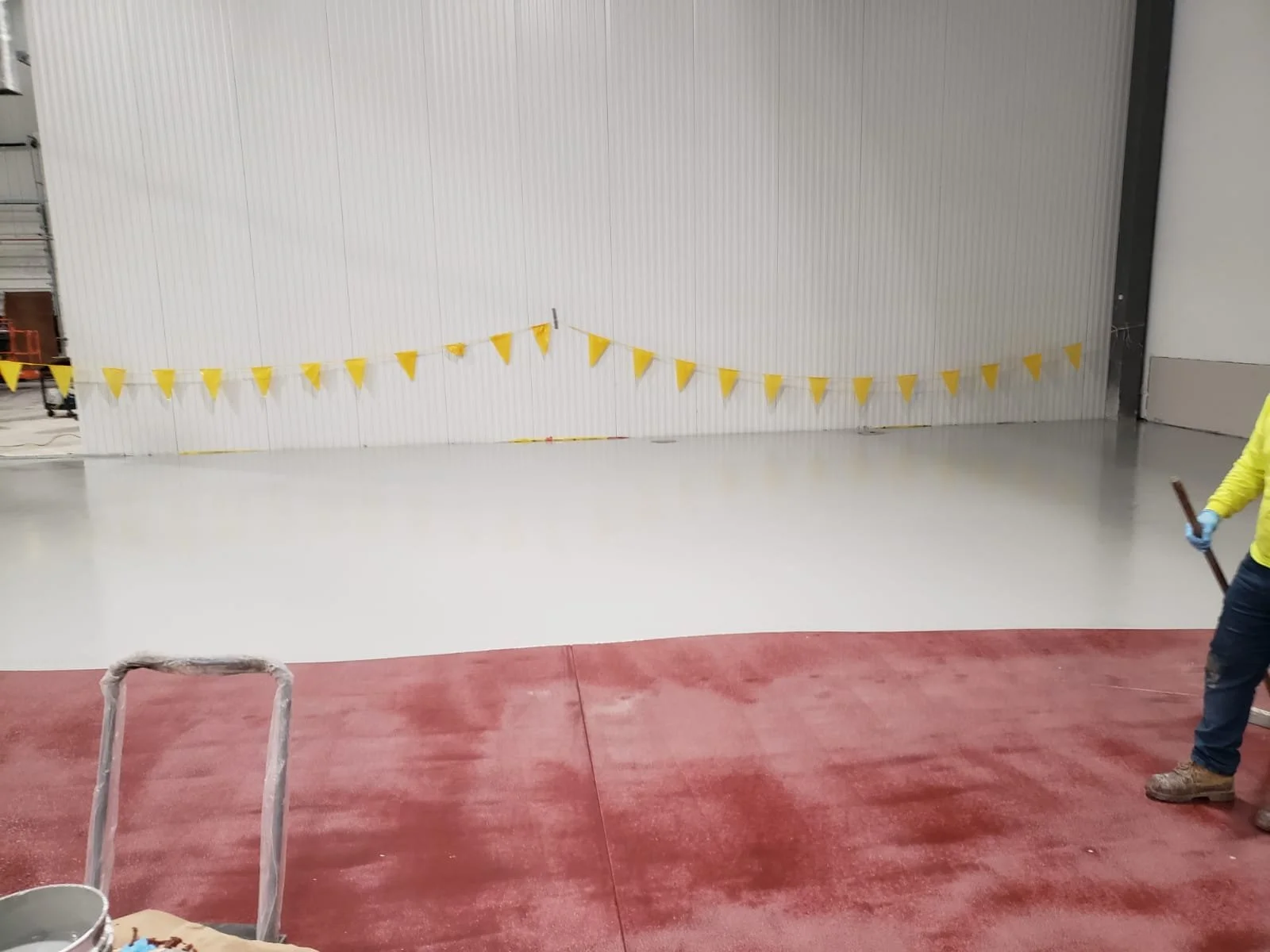 Interior view of a building under construction, showing a partially completed red and gray concrete floor and a large white wall with yellow safety flags strung across it. A worker wearing a yellow shirt, jeans, and work boots is holding a tool on th