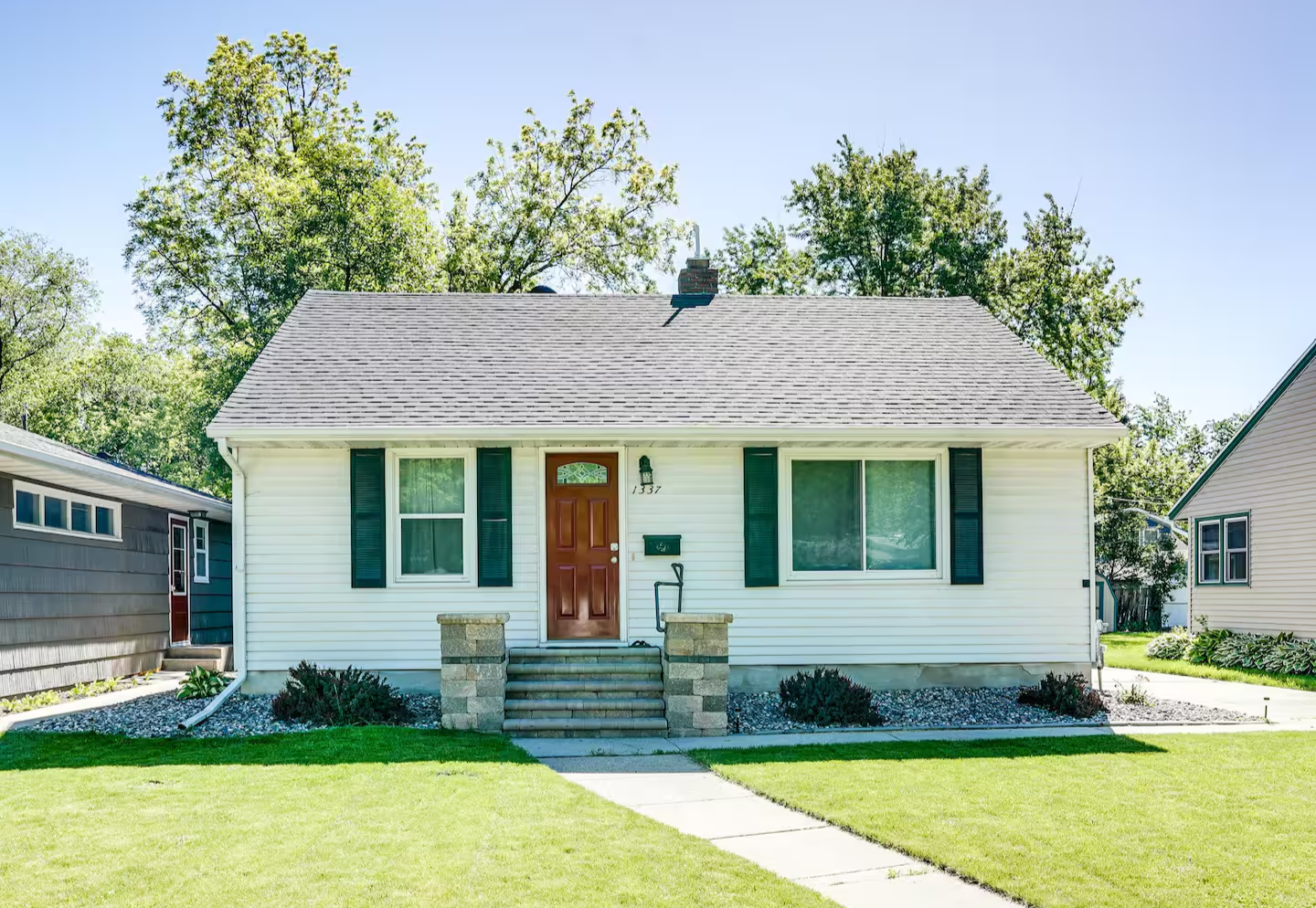 Front view of a small white house with black shutters, a brown front door, and a front porch with stone steps, surrounded by a green lawn and trees.
