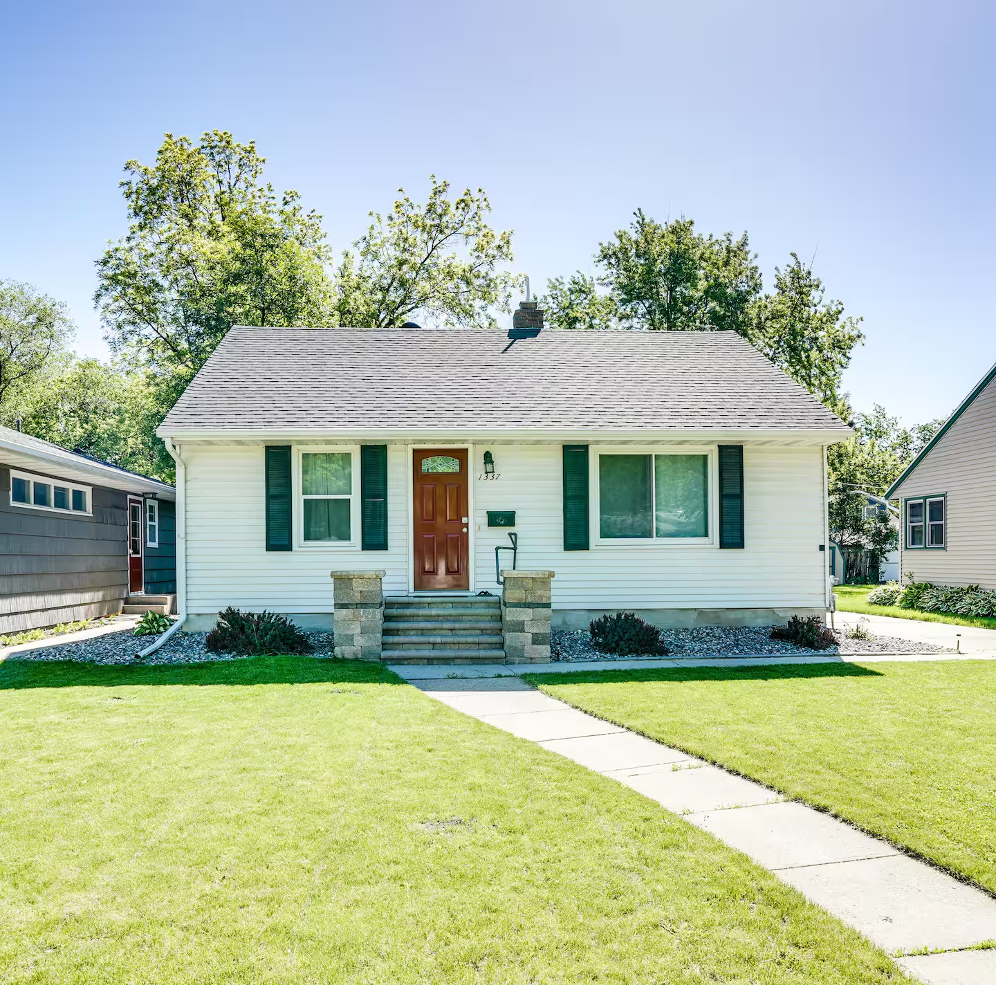 Front view of a white house with black shutters, a red front door, and a small front porch, surrounded by a green lawn and trees.