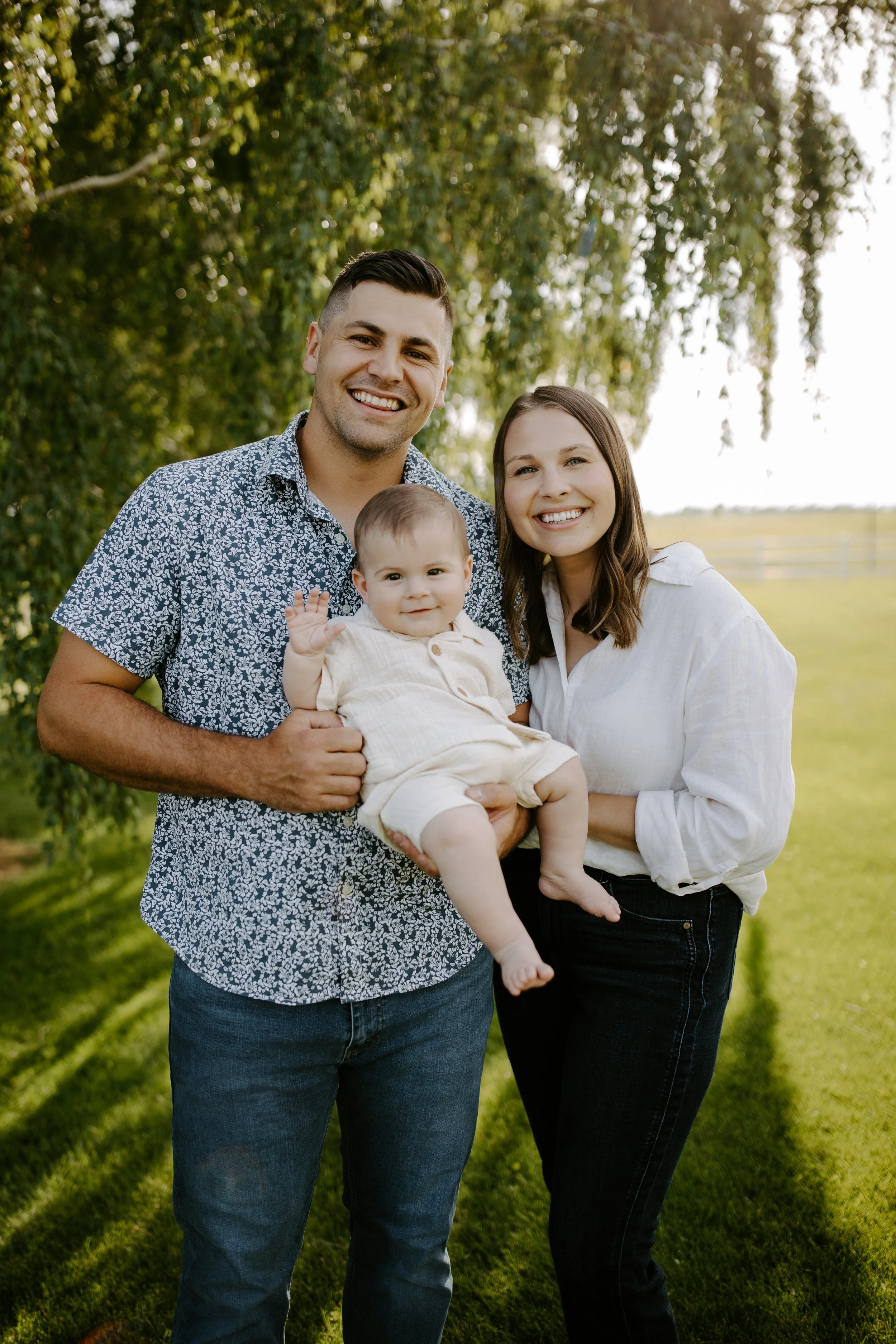 A happy family of three outdoors under a tree, smiling at the camera. The man is holding a smiling baby boy, and the woman is standing beside them in a white shirt. The background features a lush green landscape.