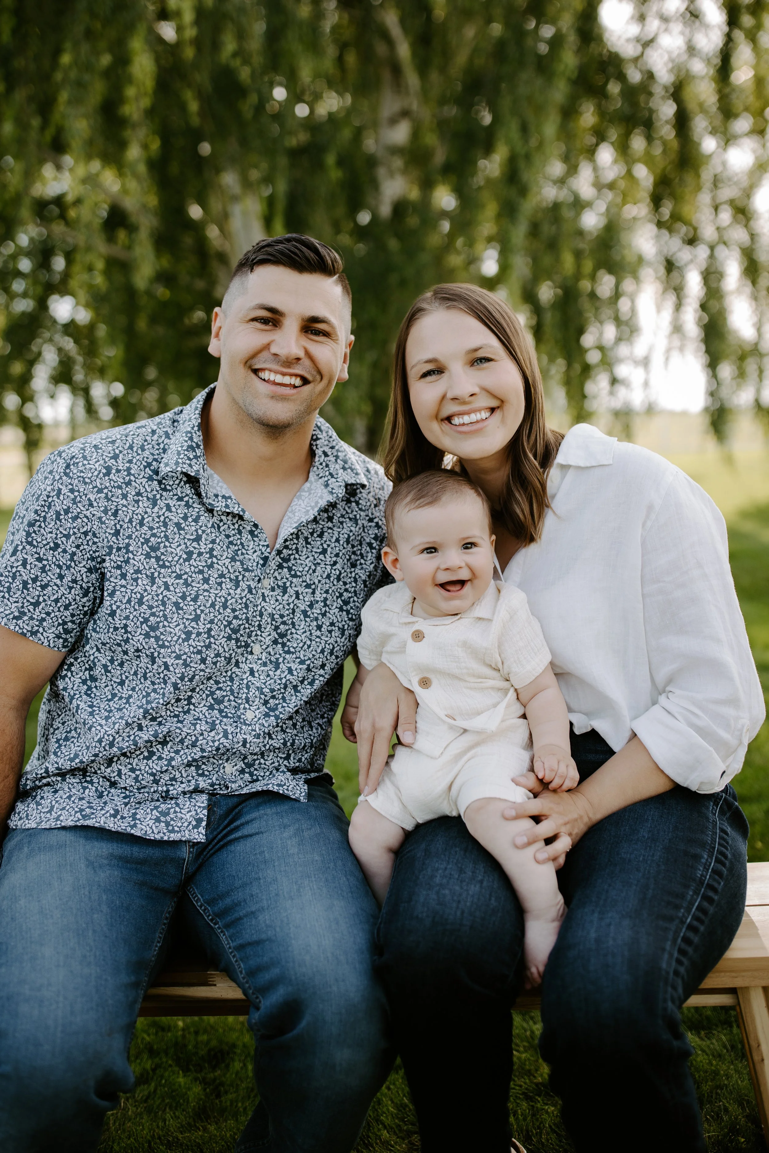 A smiling family of three sitting on a wooden bench outdoors, with green trees in the background. The father and mother are smiling and holding their baby, who is also smiling, all dressed casually.