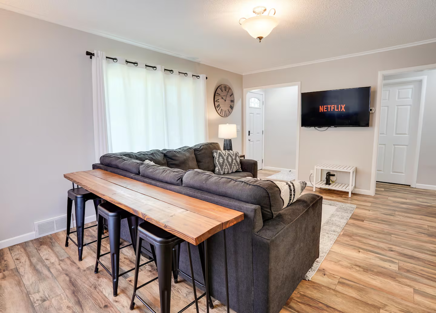 Living room with a dark gray sectional sofa, a wooden dining table with three black barstools, a wall clock, a floor lamp, a TV displaying Netflix, and a door leading to the front door. Wood flooring and white curtains on the window.