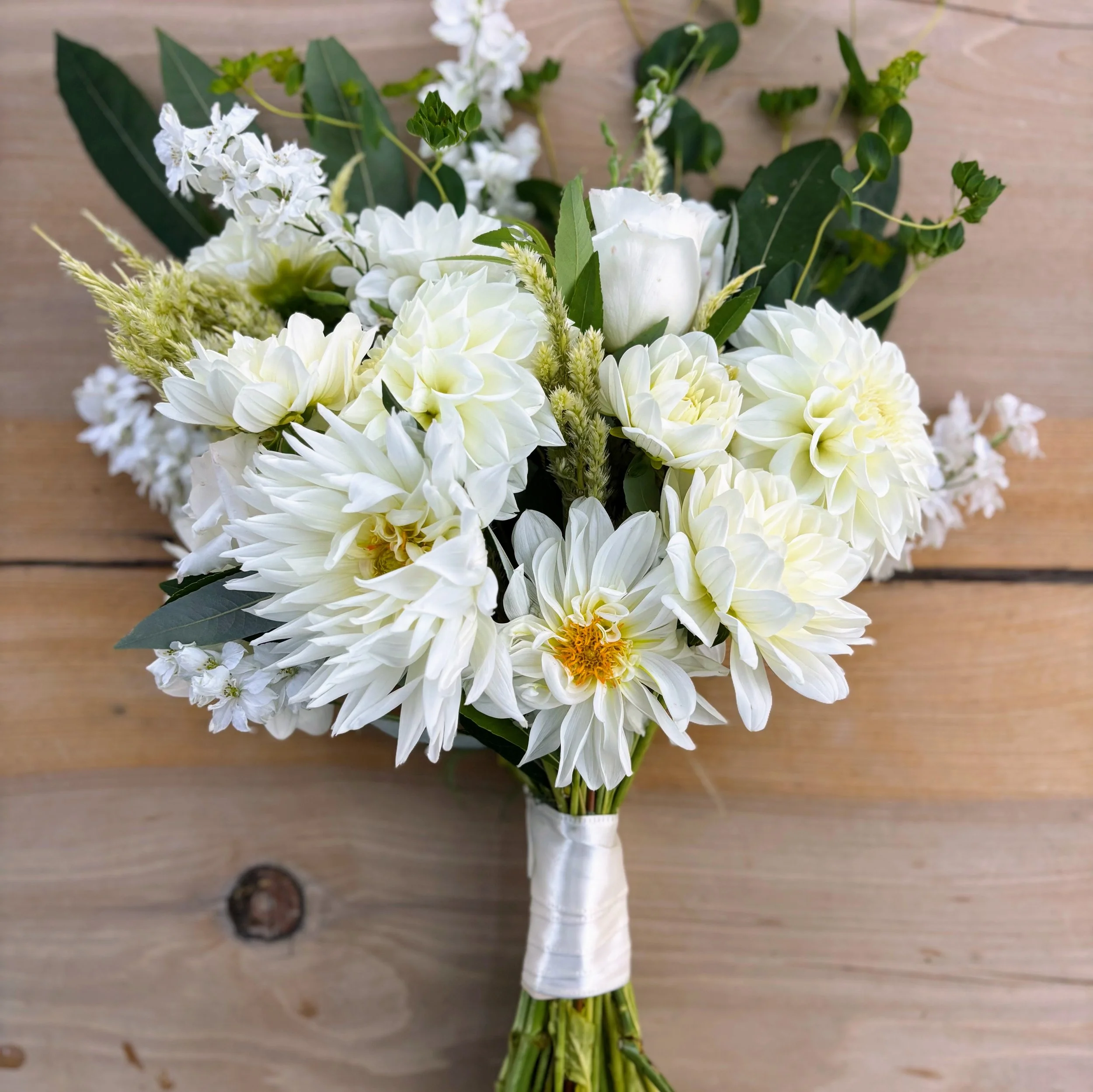 A bouquet of white flowers on a wooden surface, featuring large white dahlias, roses, and green leaves.