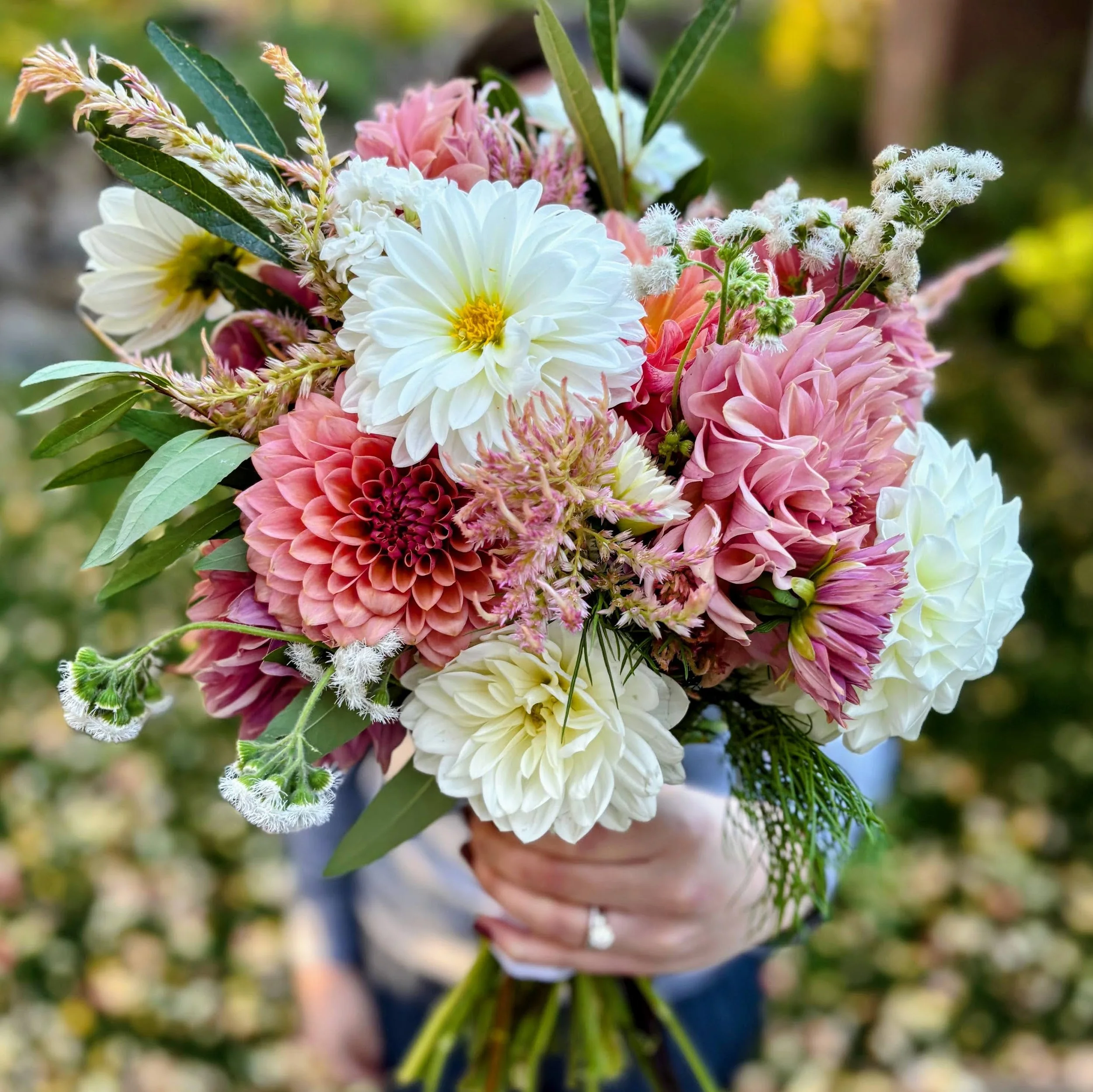 A person holding a colorful bouquet of pink, white, and cream flowers with green leaves.