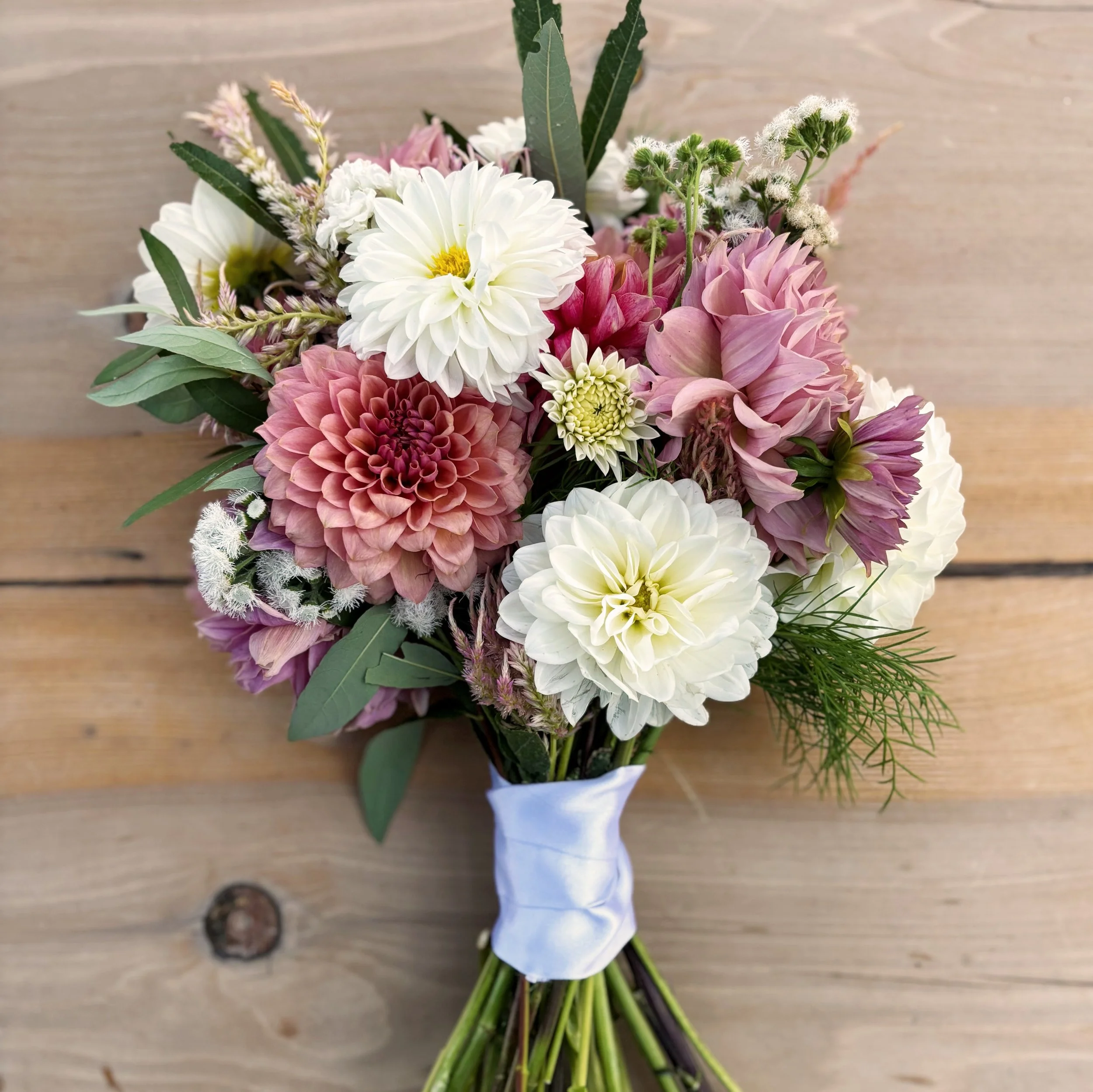 A bouquet of various pink and white flowers with green foliage, tied with a white ribbon, placed on a wooden surface.