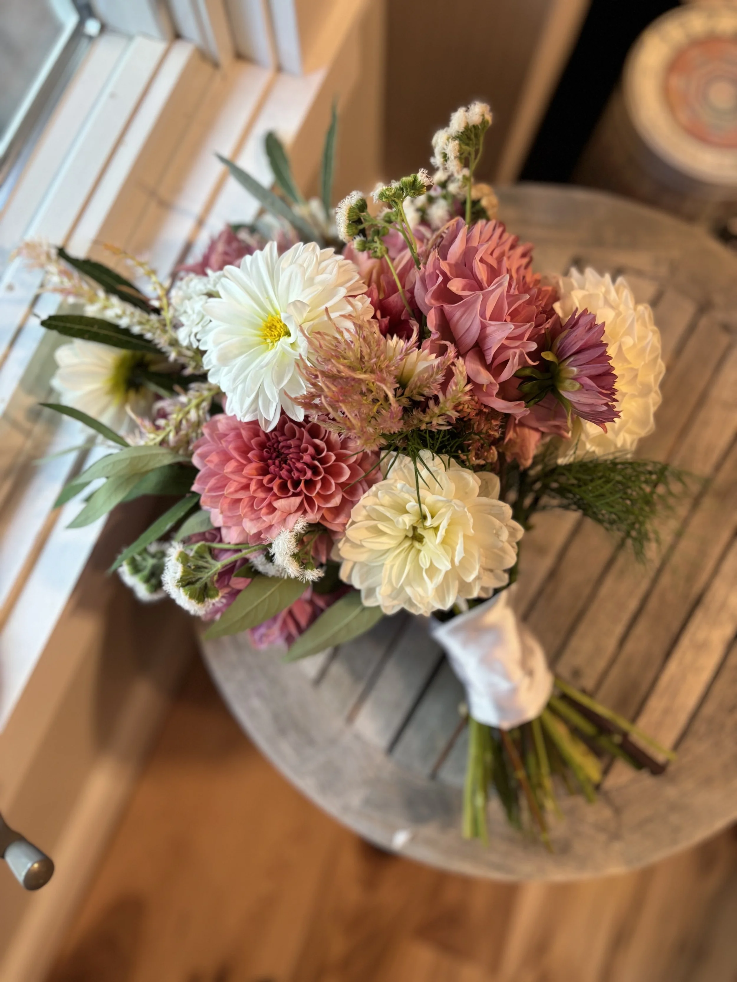 A colorful bouquet of pink, white, and cream flowers with green leaves in a black vase, sitting on a round wooden table.