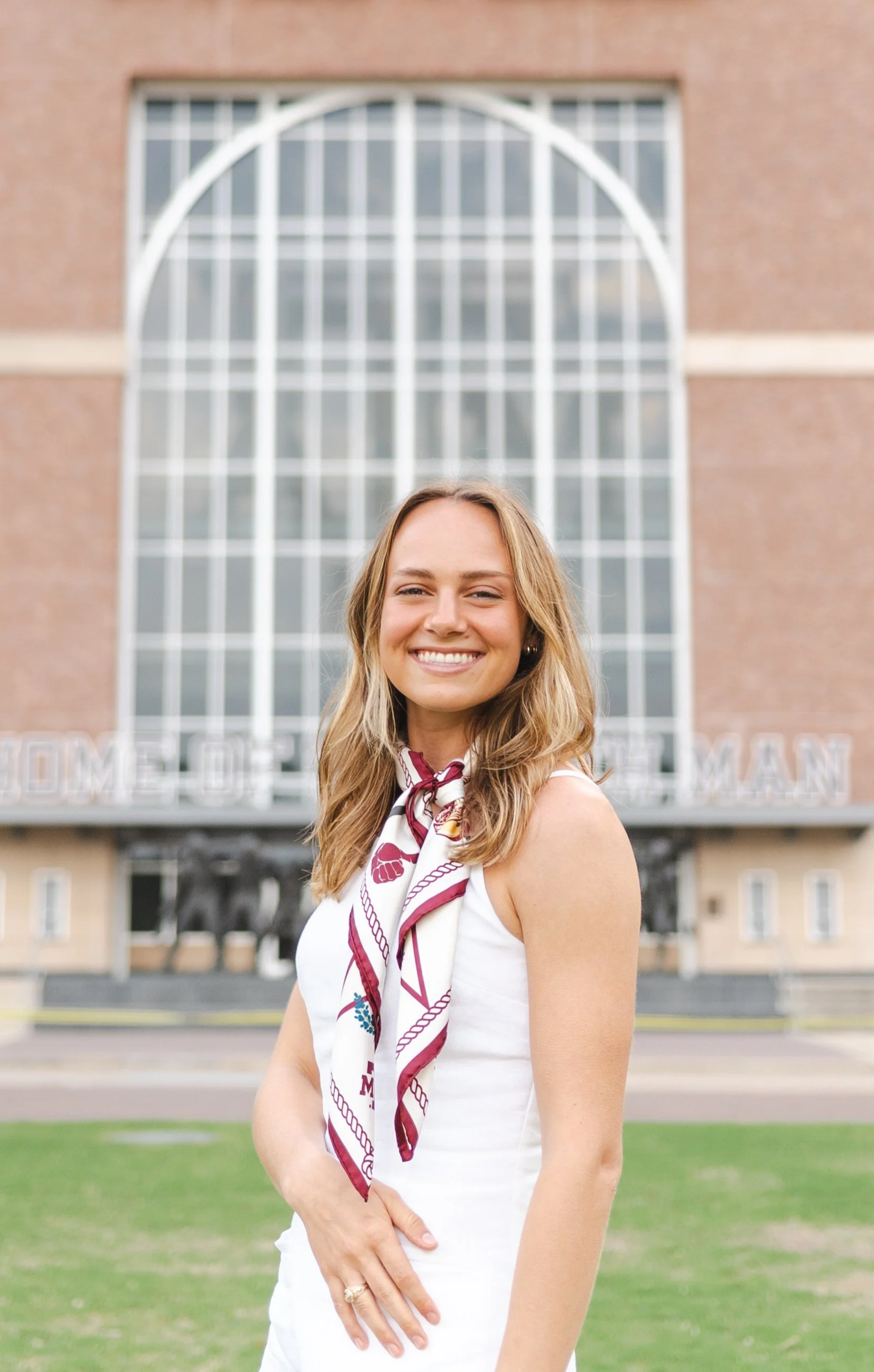 A young woman with long wavy blonde hair, smiling, wearing a white sleeveless dress and a maroon and white neck scarf, standing outdoors in front of a large brick building with tall windows.