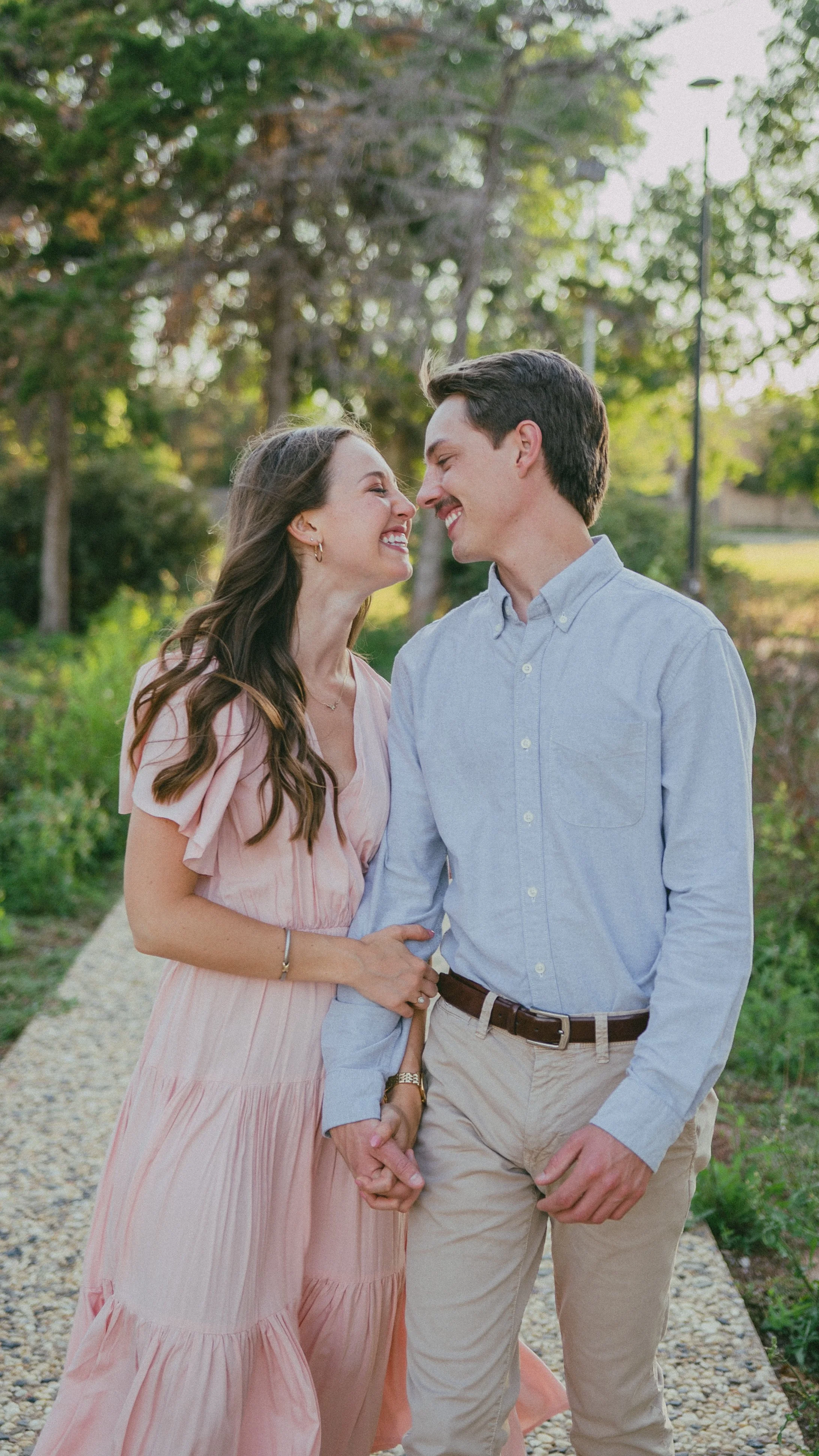A happy couple holding hands and smiling at each other outdoors on a sunny day, with trees and greenery in the background.