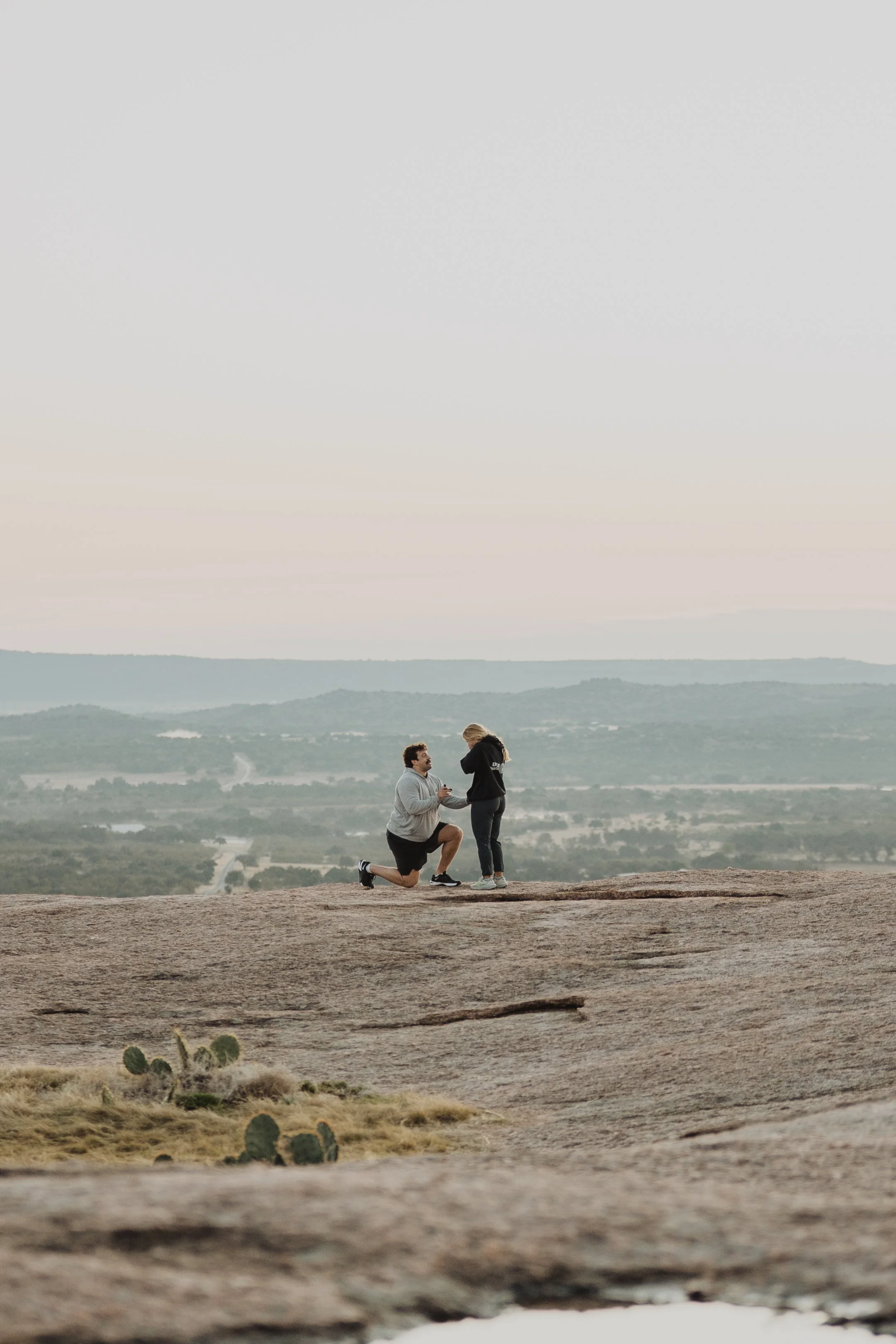 A man proposing marriage to a woman on a rocky hilltop during sunset with a view of a valley and distant mountains.