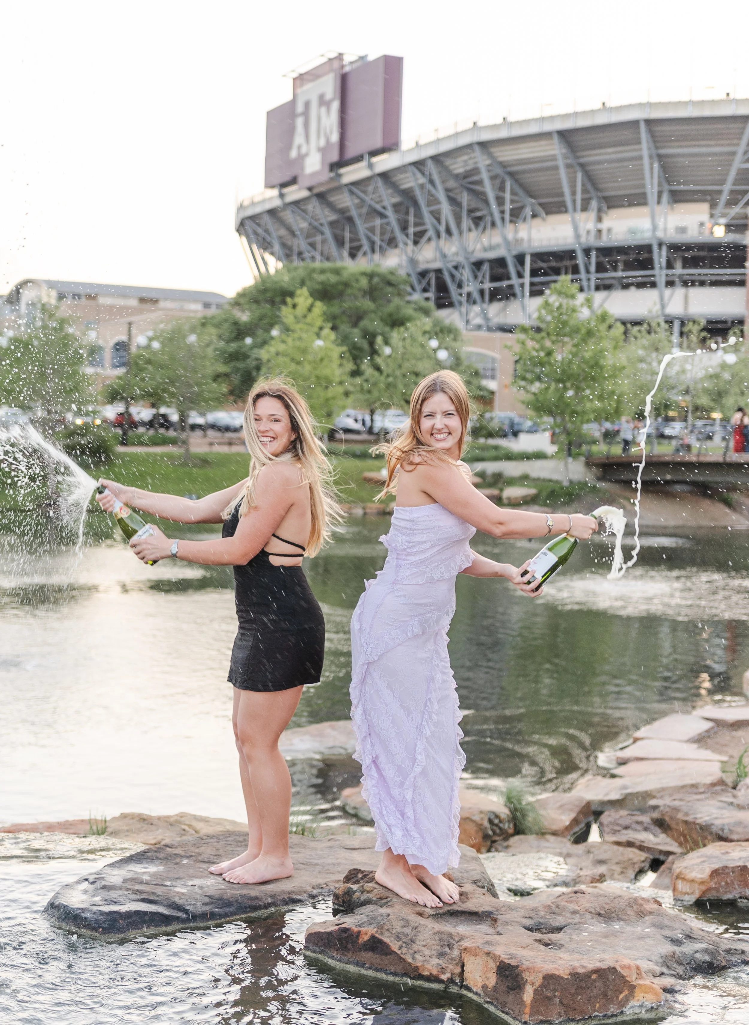 Two young women in dresses celebrating on rocks near a pond, spraying champagne, with a stadium in the background.