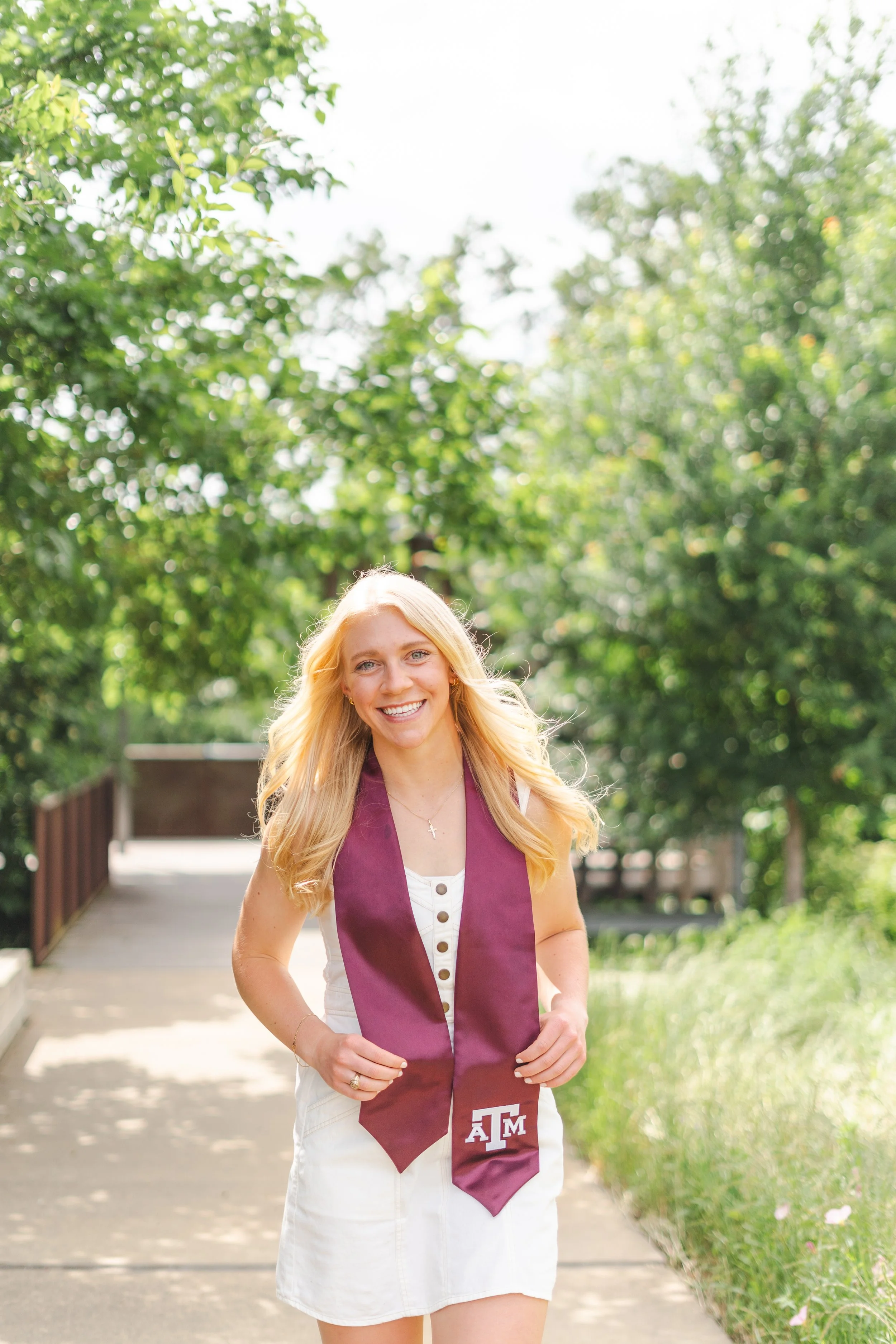 A young woman with blonde hair smiling and walking outdoors on a sunny day, wearing a white dress and a maroon graduation stole with the Texas A&M logo.