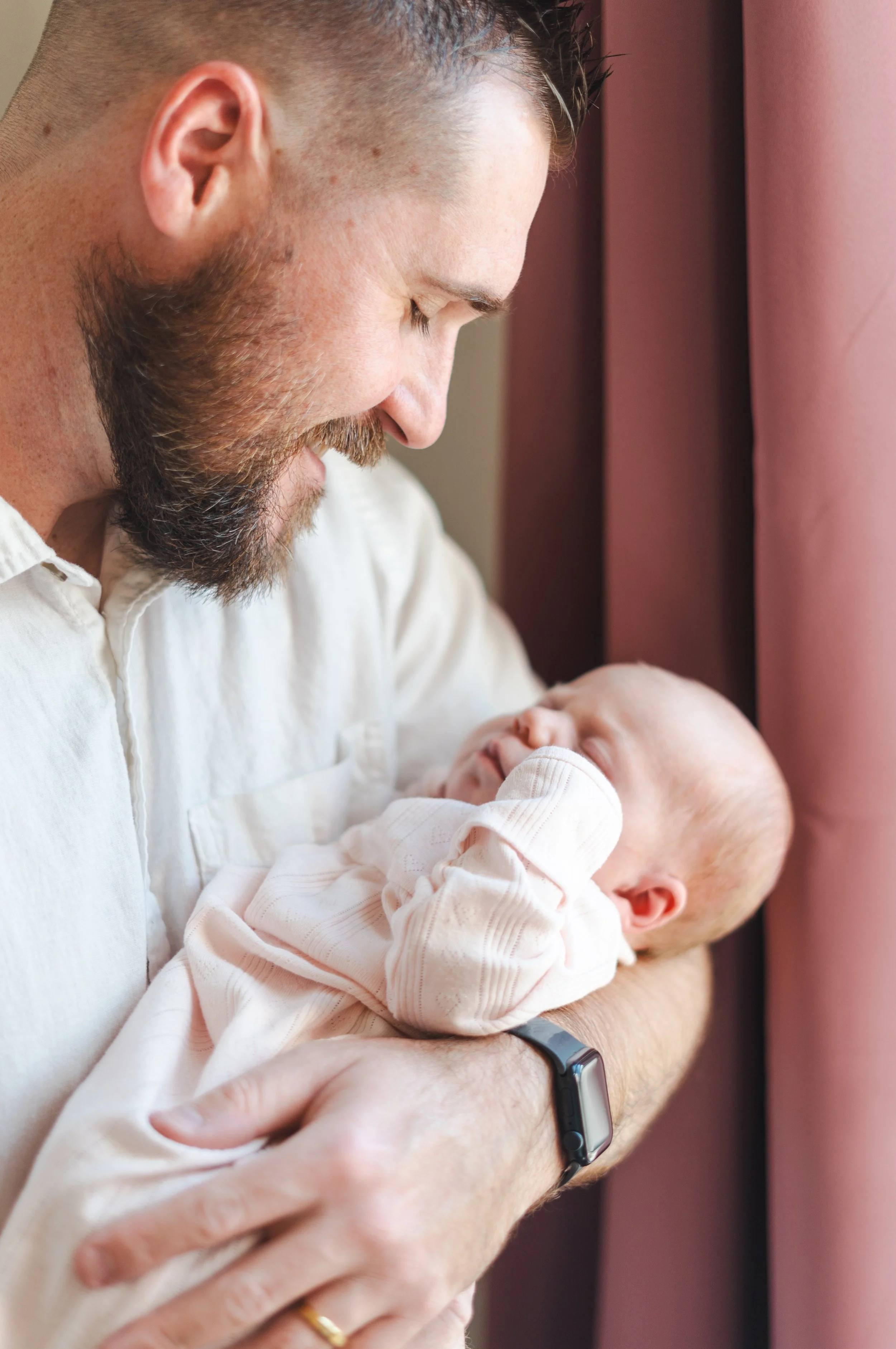A man with a beard holds a sleeping baby close to his chest, smiling softly while looking at the baby, near a pink curtain.