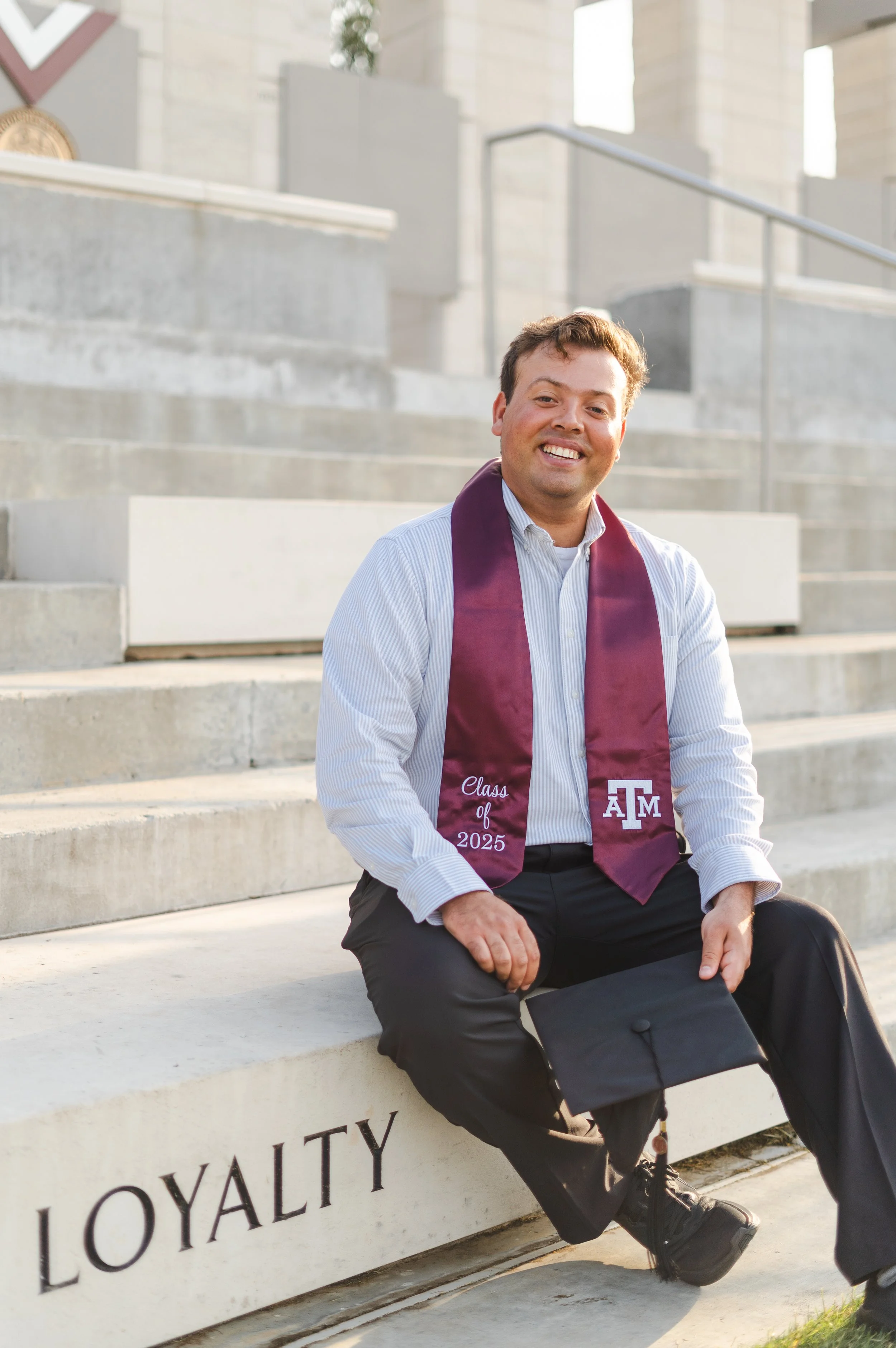 A young man in formal attire sitting on concrete steps, wearing a white dress shirt, black pants, and a maroon graduation stole with 'Class of 2025' and 'TAMU' embroidered on it, holding a graduation cap with a smile.