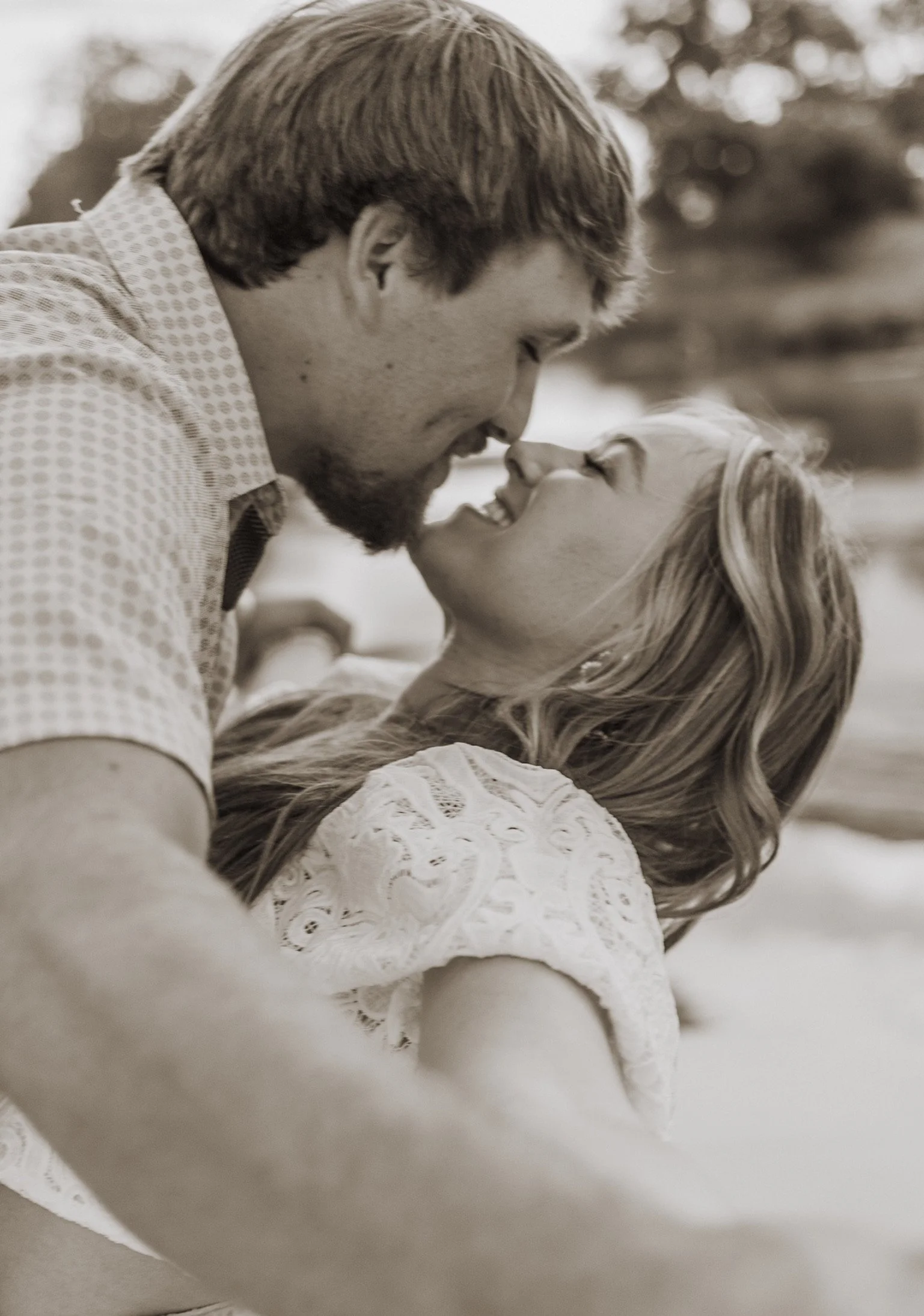 A man and woman sharing a close, intimate moment outdoors, smiling and leaning towards each other.