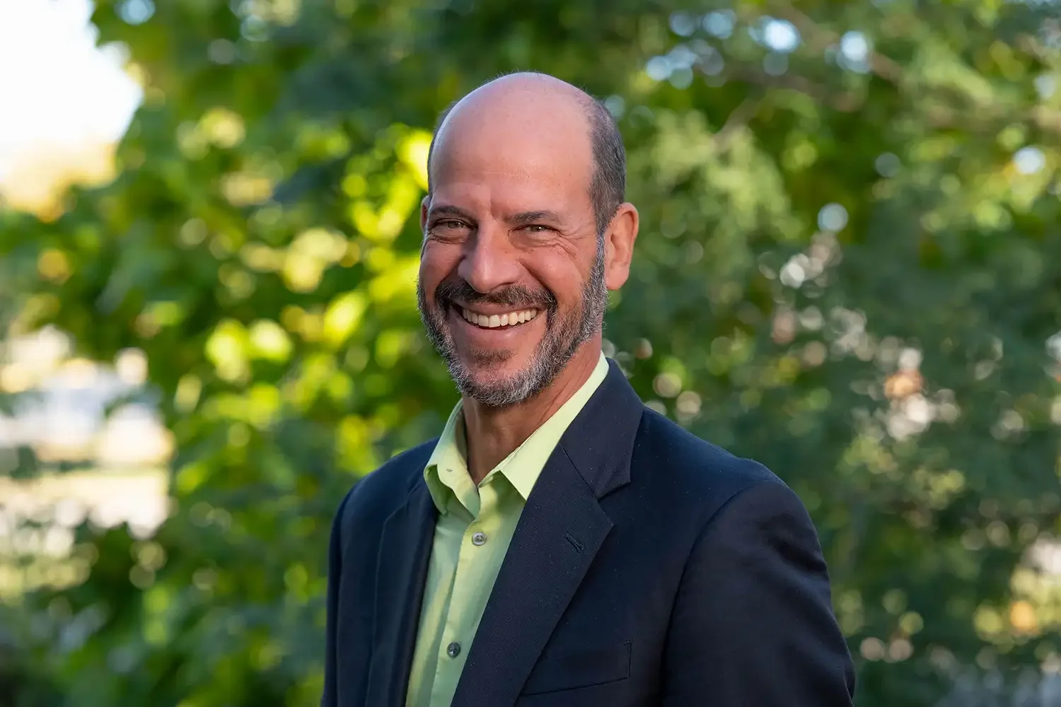 A smiling middle-aged man with a beard and no hair, wearing a black blazer and light green shirt, standing outdoors with a blurred green leafy background.