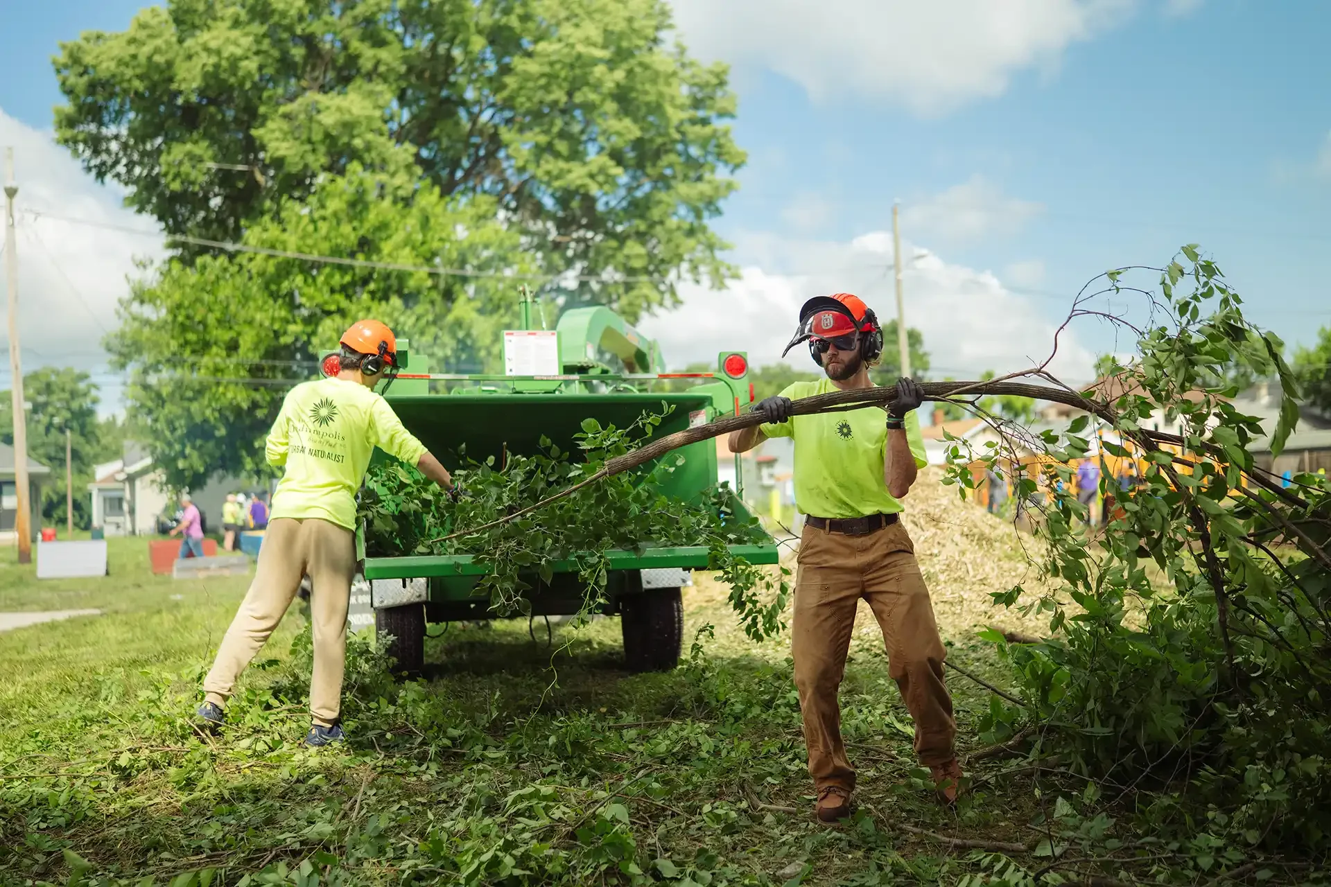 Two workers wearing yellow shirts, helmets, and safety gear are clearing fallen branches into a green trash truck in a park during daytime. One worker is lifting a large branch, while the other is placing branches into the truck.
