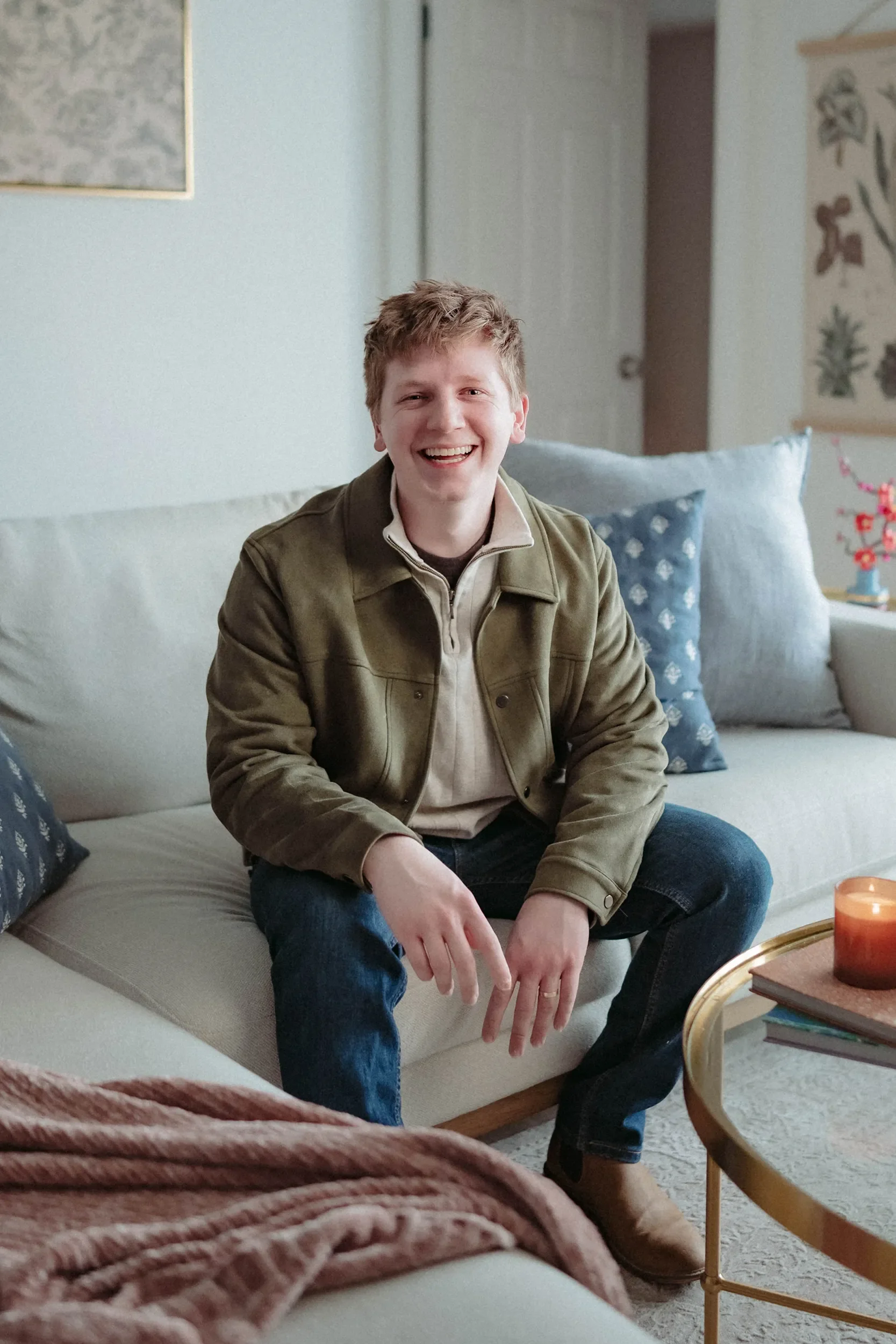 A young man with curly blonde hair smiling and sitting on a beige sofa in a cozy living room.