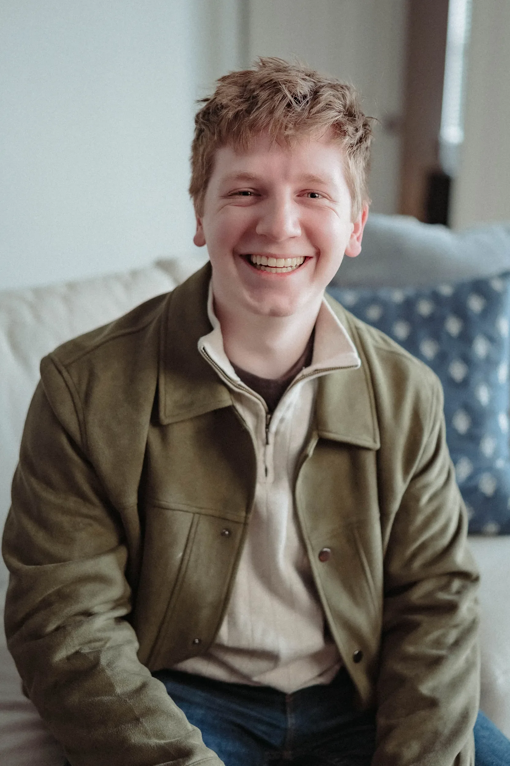 A young man with curly hair smiling widely, seated on a sofa with a cream-colored pillow and a blue patterned pillow behind him, wearing a tan jacket and cream zipper pullover.