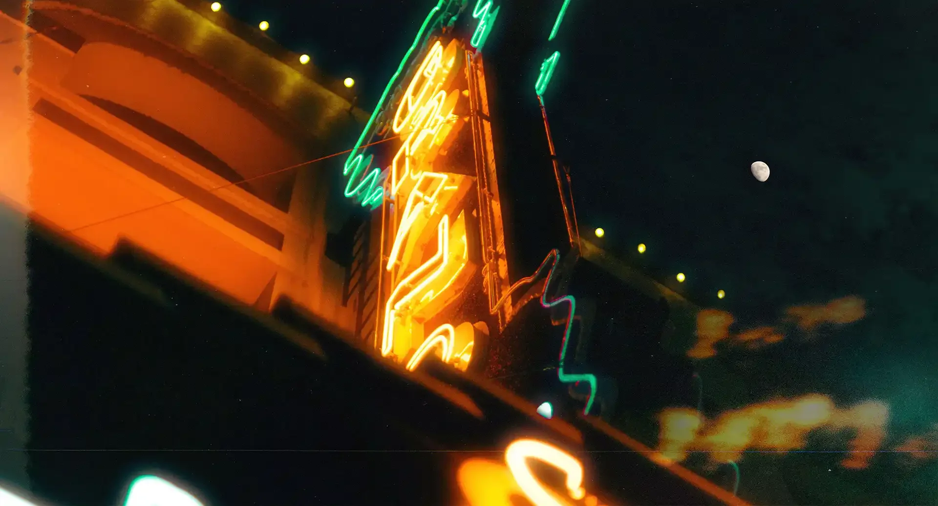 Neon signs glowing in orange and green against a dark night sky with the moon visible.