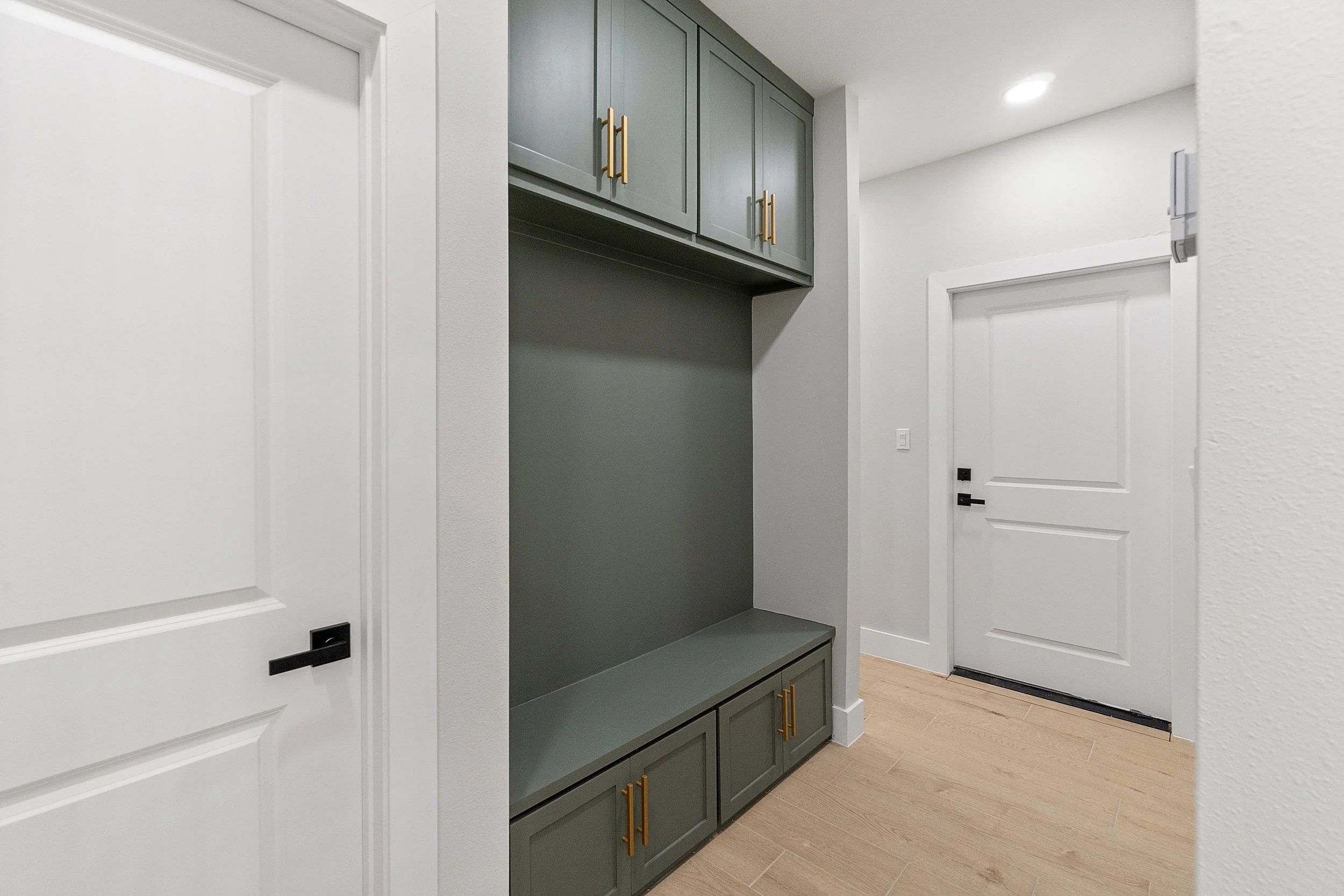 Entryway with a light green built-in bench and cabinet with gold handles, white walls, light wooden flooring, and two white doors, one with black hardware.