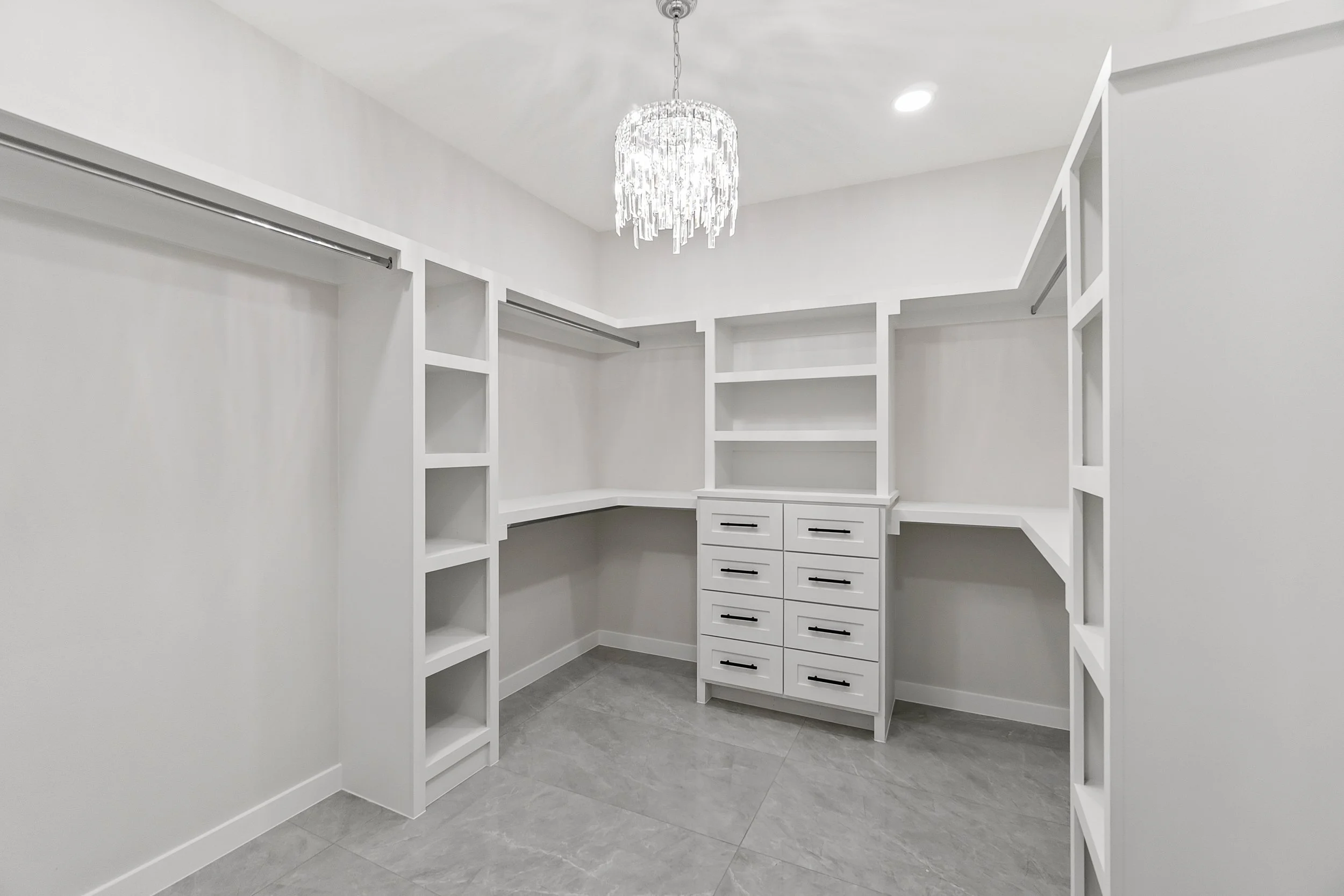 Empty white walk-in closet with built-in shelves, drawers, and hanging rods, modern chandelier, gray floor tiles, and white walls.