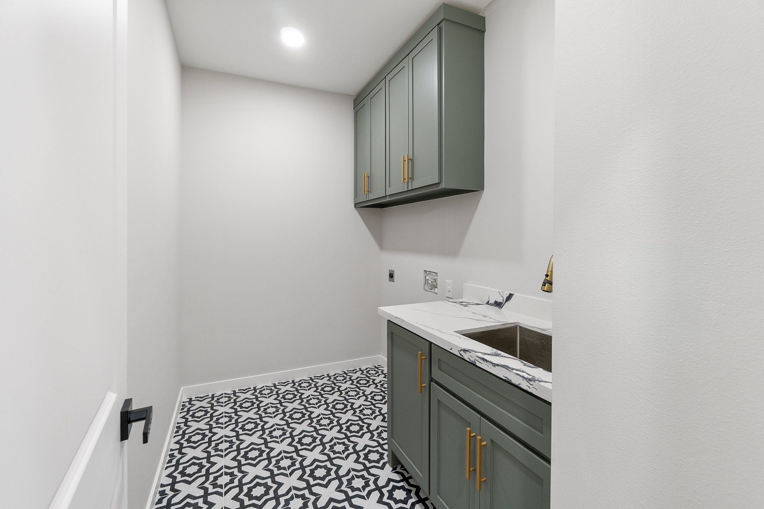 Small laundry room with grey cabinets, marble countertop, and patterned black and white tile floor.