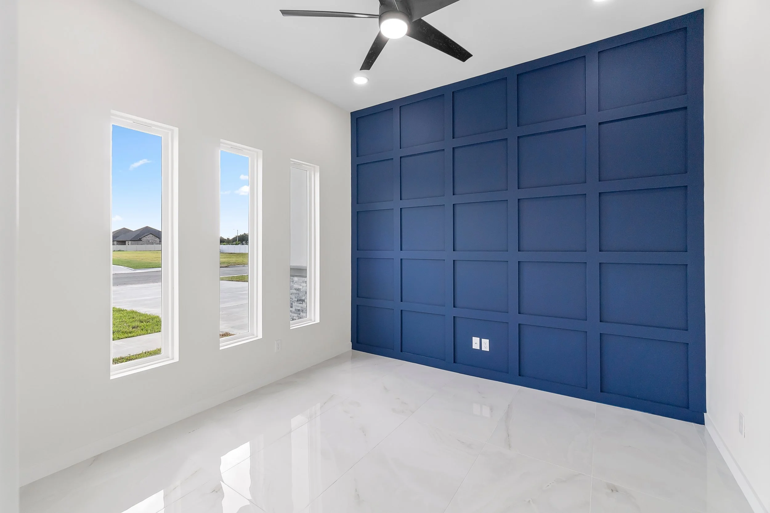 Empty room with white walls, three tall narrow windows, a blue accent wall with a grid pattern, a ceiling fan, and glossy white floor tiles.
