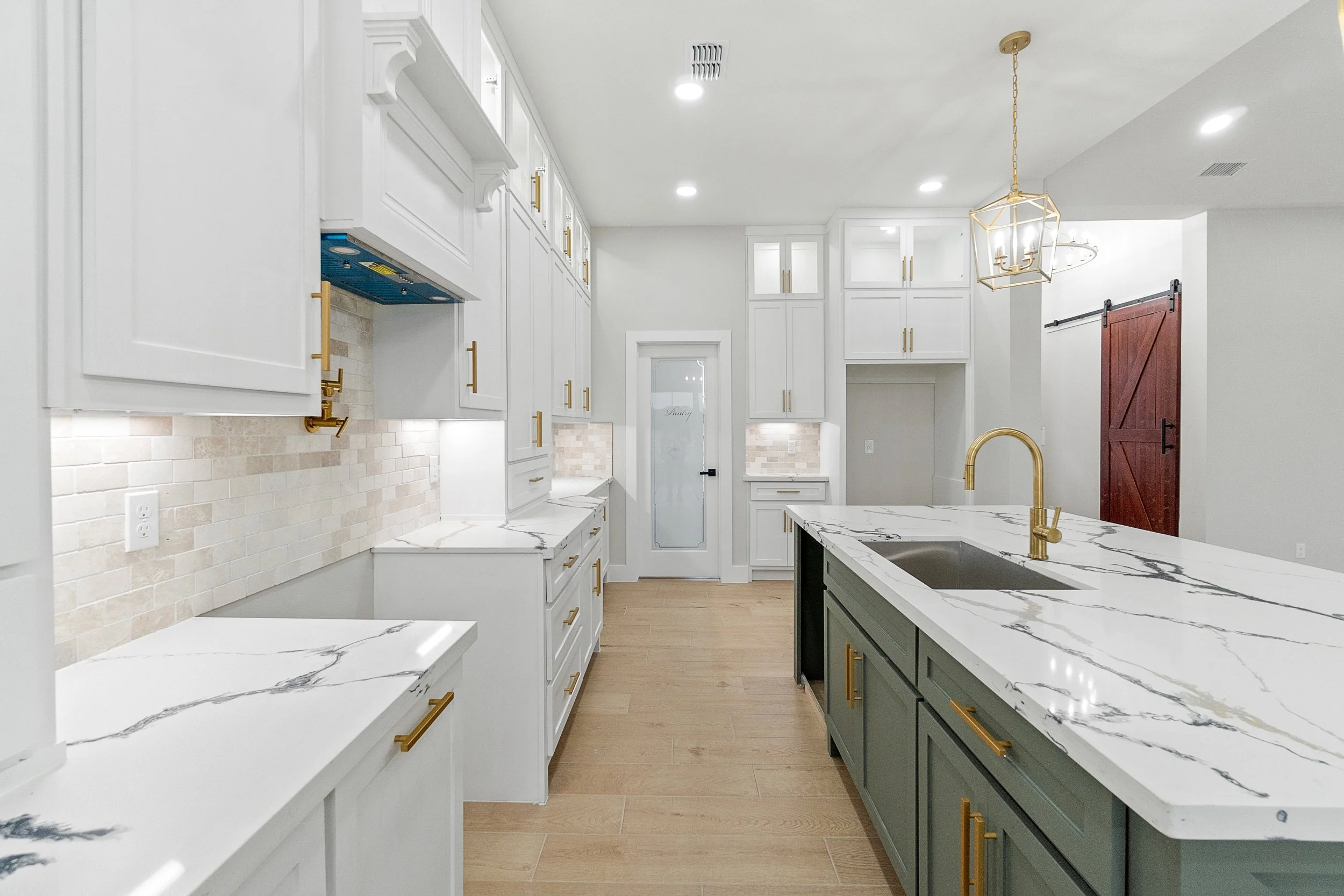 Modern kitchen with white cabinetry and gold hardware, a marble island with a gold faucet, beige brick backsplash, and light wood flooring.