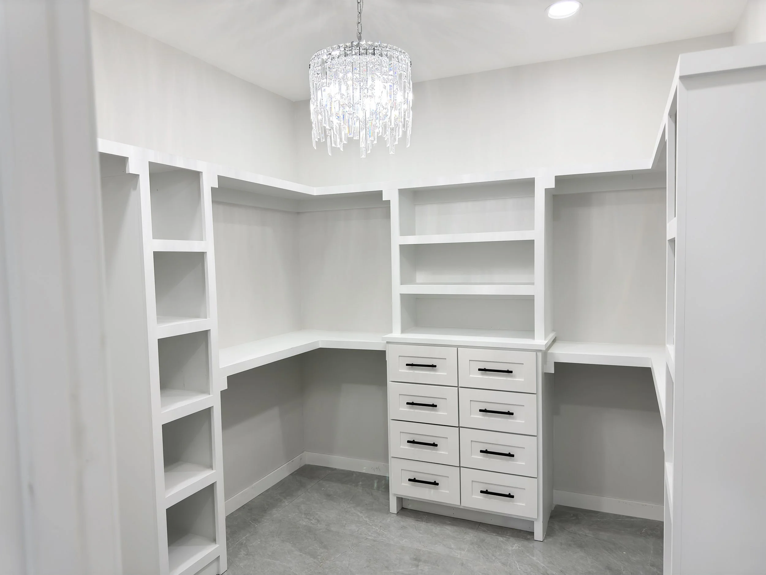 Empty white walk-in closet with built-in shelving, drawers, and a crystal chandelier