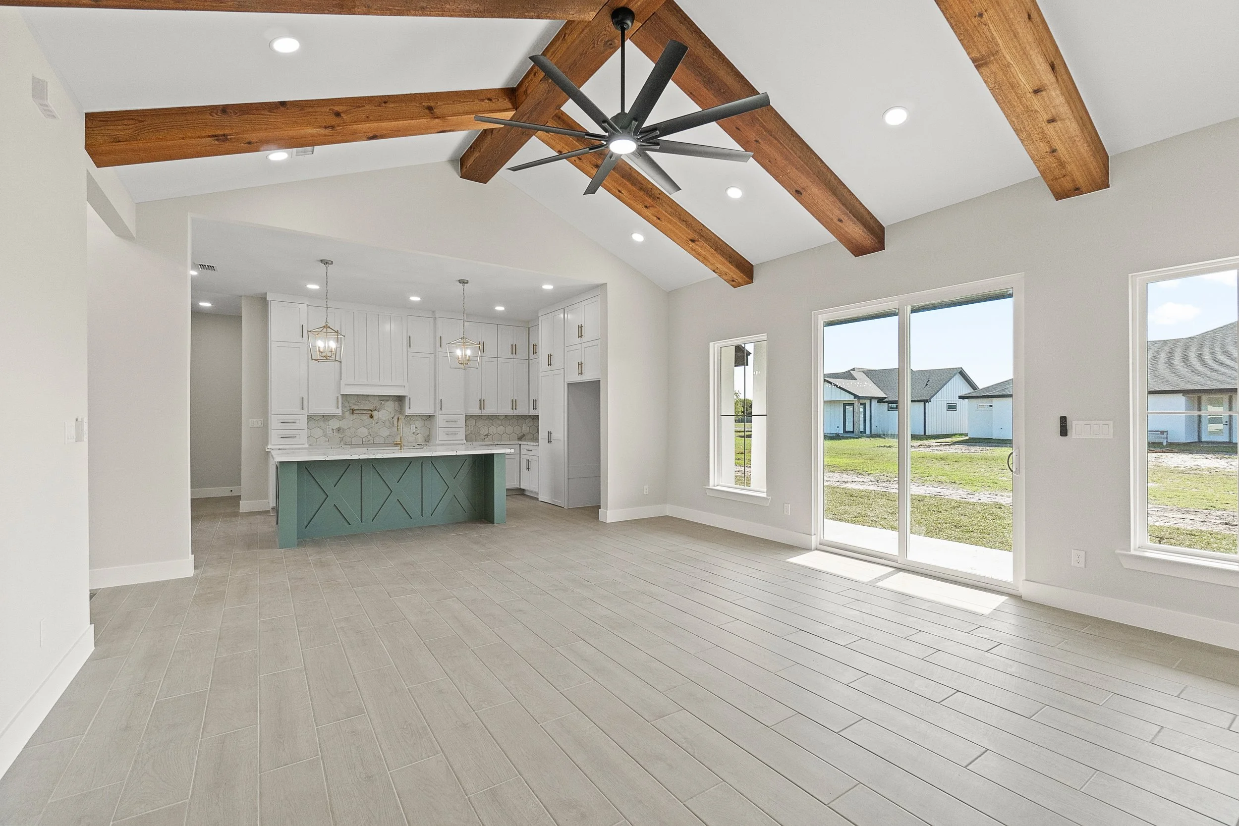Empty living room with large windows, sliding glass door, and high vaulted ceiling with wooden beams and a ceiling fan.