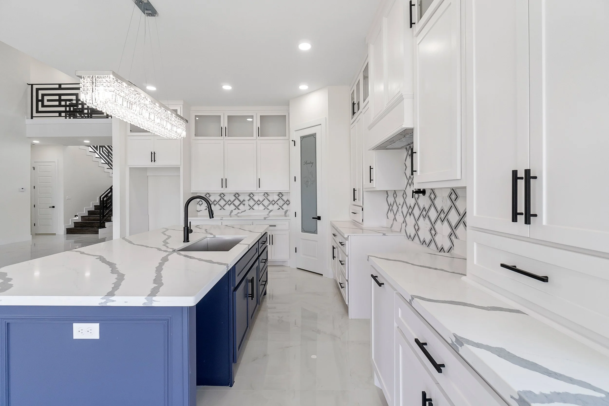 Modern kitchen with white cabinets, black handles, marble countertops with gray veining, and a blue kitchen island. Recessed lighting and a rectangular chandelier with crystals above the island. A hallway with stairs is visible in the background.