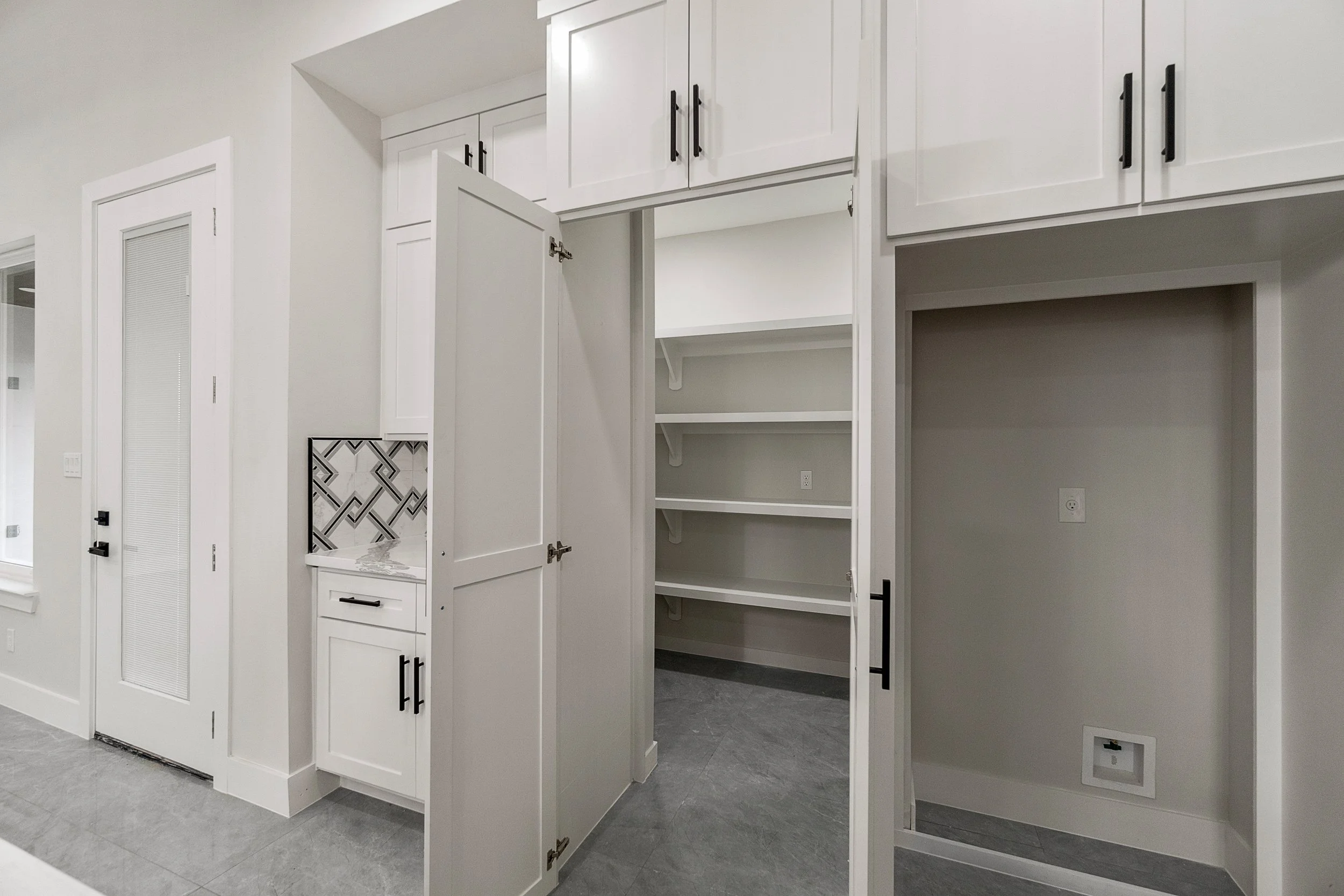Empty white pantry with open doors, multiple shelves, and a nearby laundry area with a blank wall and an electrical outlet.