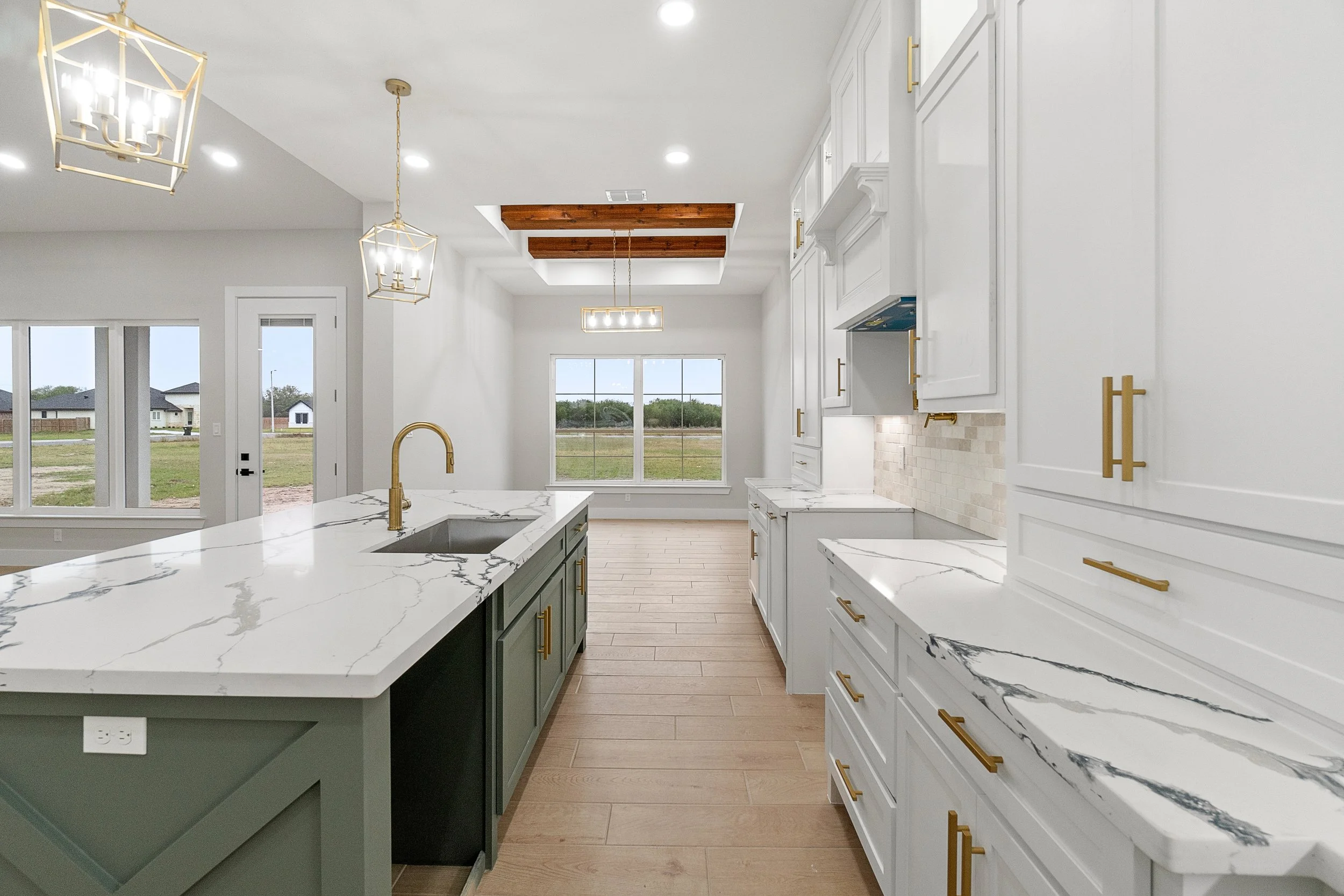 Modern kitchen with white and green cabinetry, marble countertops, gold handles, a central island with a sink, and large windows providing natural light.