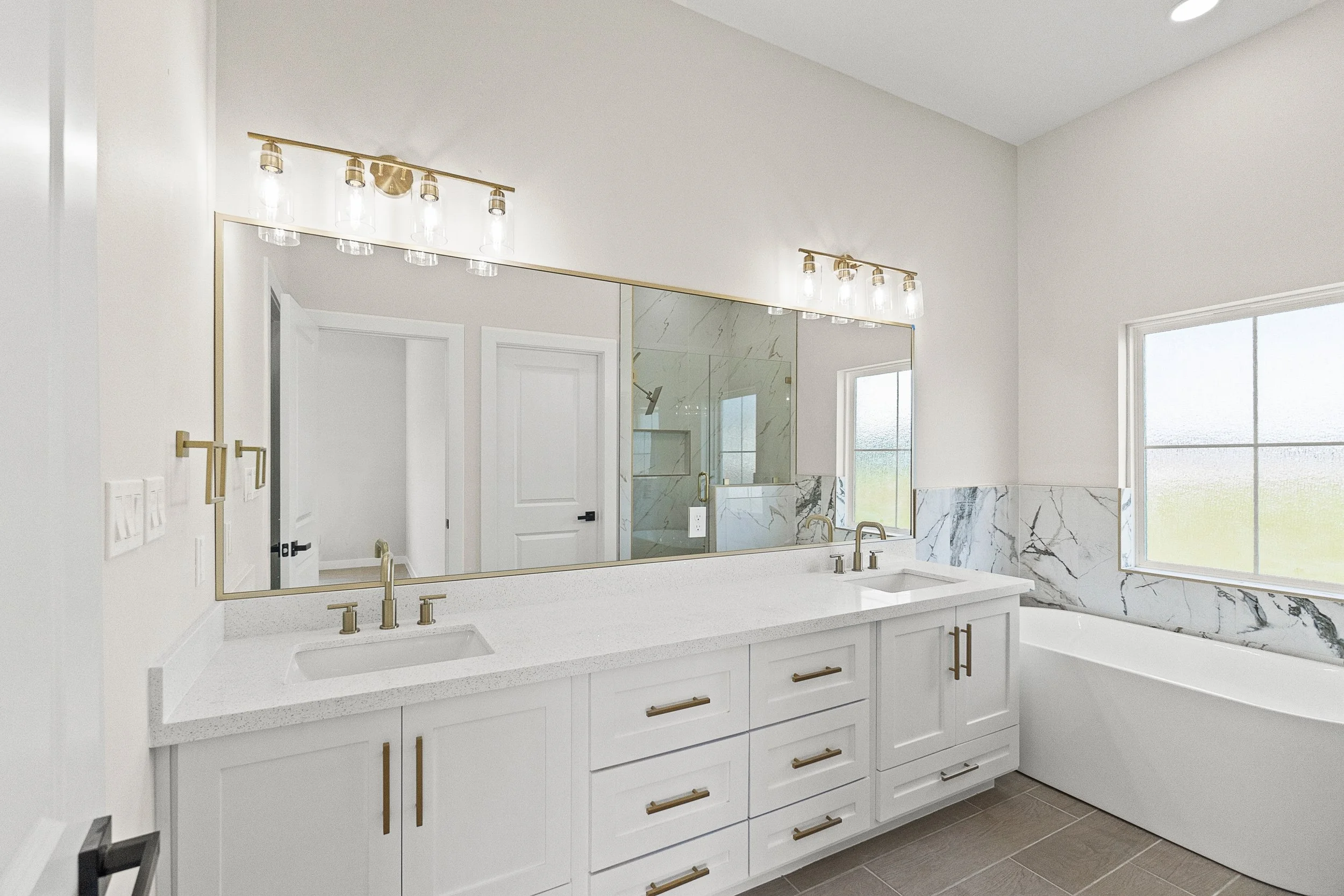 Modern bathroom with white vanity, dual sinks, gold hardware, large mirror, marble accents, bathtub, and windows