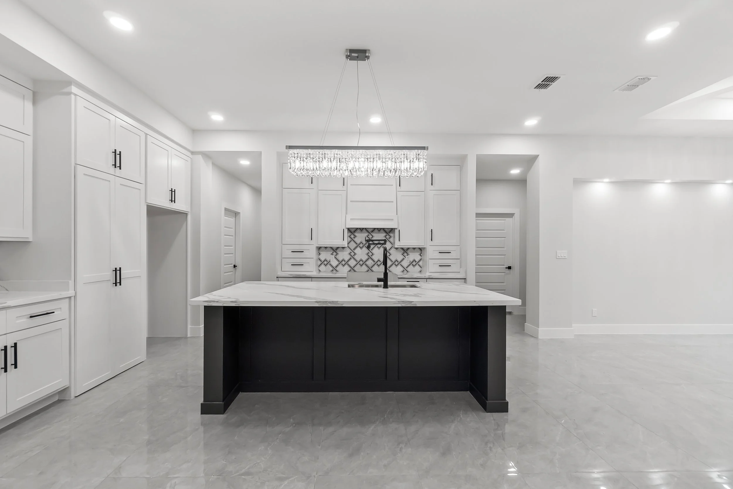 A modern, spacious kitchen with white cabinets, an island with a black base and marble countertop, a pendant chandelier, and a patterned backsplash.