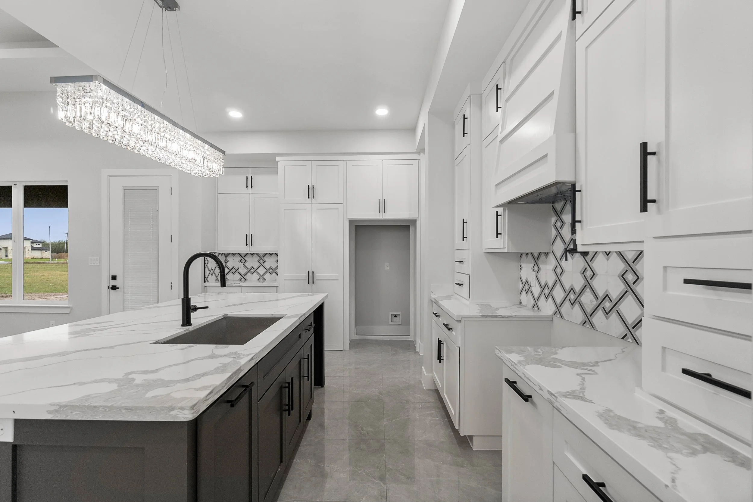 Modern kitchen with white cabinets, a large island with a marble countertop, black hardware, and a patterned backsplash. Recessed lighting and a chandelier are also visible.